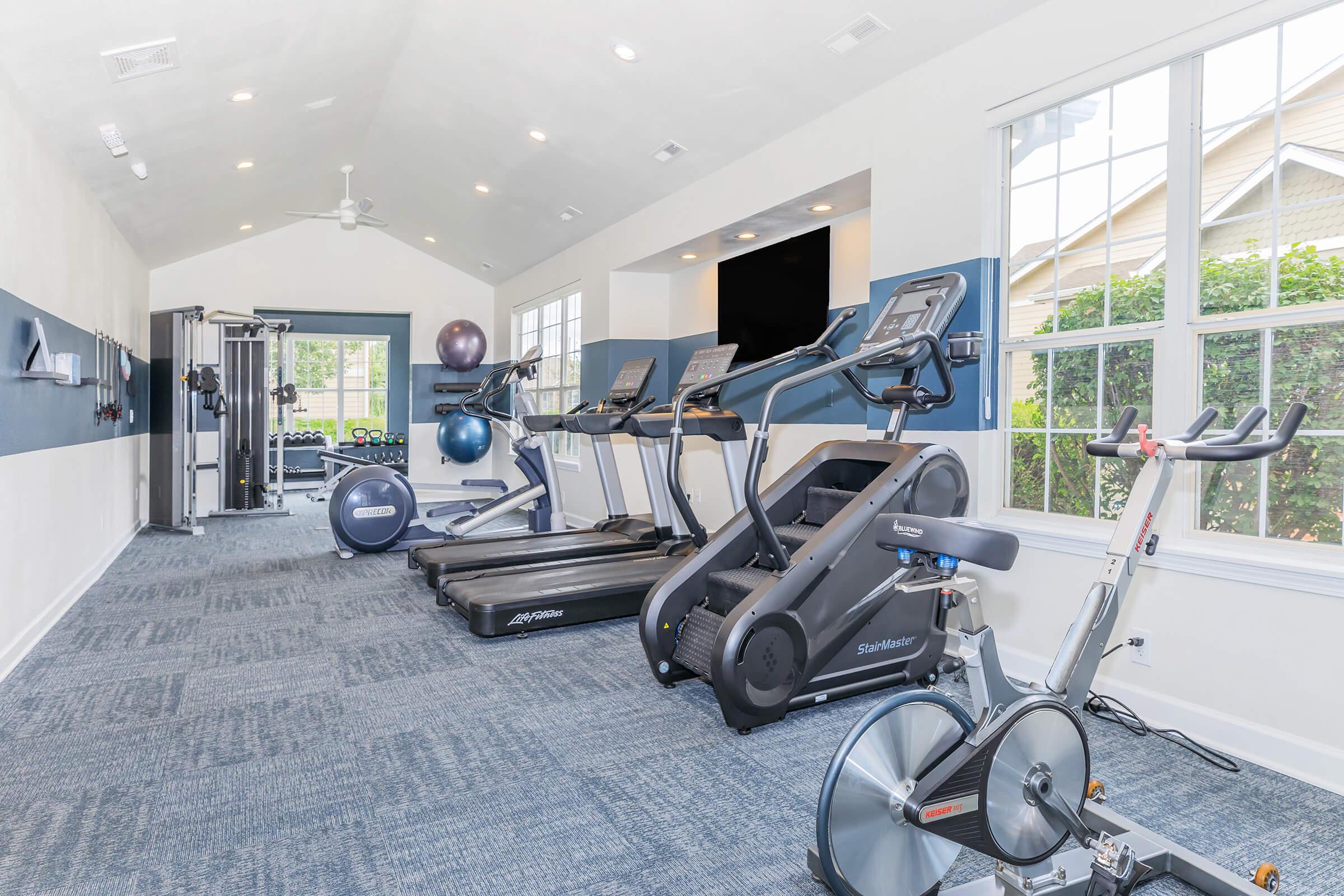 A modern fitness room featuring various exercise equipment, including treadmills, elliptical machines, and a stationary bike. Large windows provide natural light, and the walls are adorned with gym accessories. The space is tidy with a clean, contemporary design, inviting for workouts.