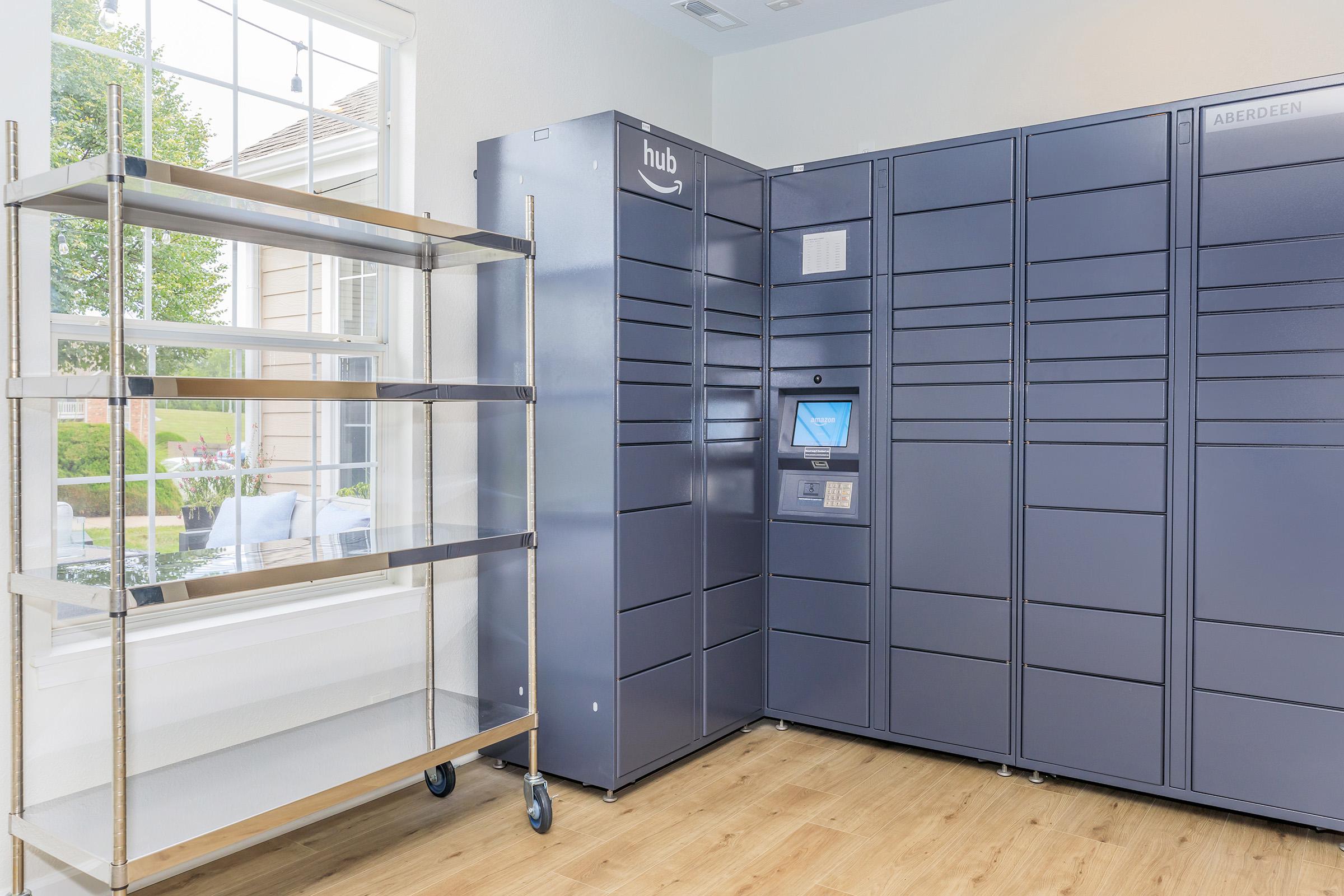 A modern package delivery locker system, featuring several grey lockers with a digital touch screen display in a well-lit room. Next to the lockers is a metal shelving unit on wheels. A window allows natural light to illuminate the space, which has wooden flooring and a view of greenery outside.