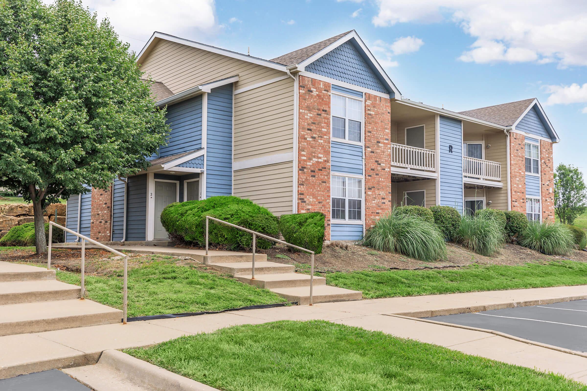 Exterior view of a two-story apartment building, featuring a mix of blue and beige siding along with brick accents. The building has balconies on the upper level, grassy landscaping, and a walkway leading up to the entrance. Additional greenery and trees are visible around the property.