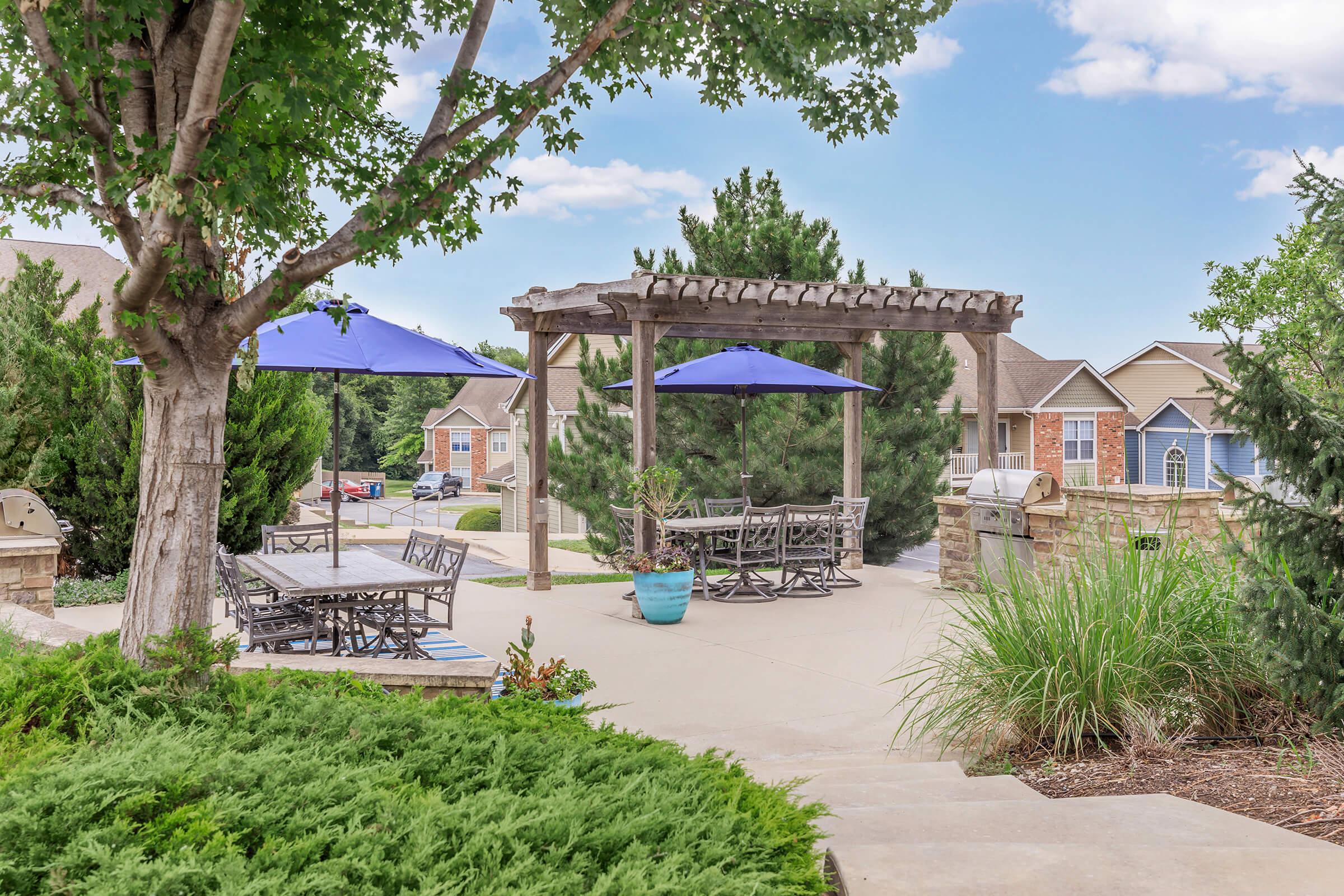 A landscaped outdoor patio area featuring a pergola with blue umbrellas, wooden tables and chairs, surrounded by trees and shrubs. In the background, residential houses are partially visible, creating a tranquil suburban atmosphere.