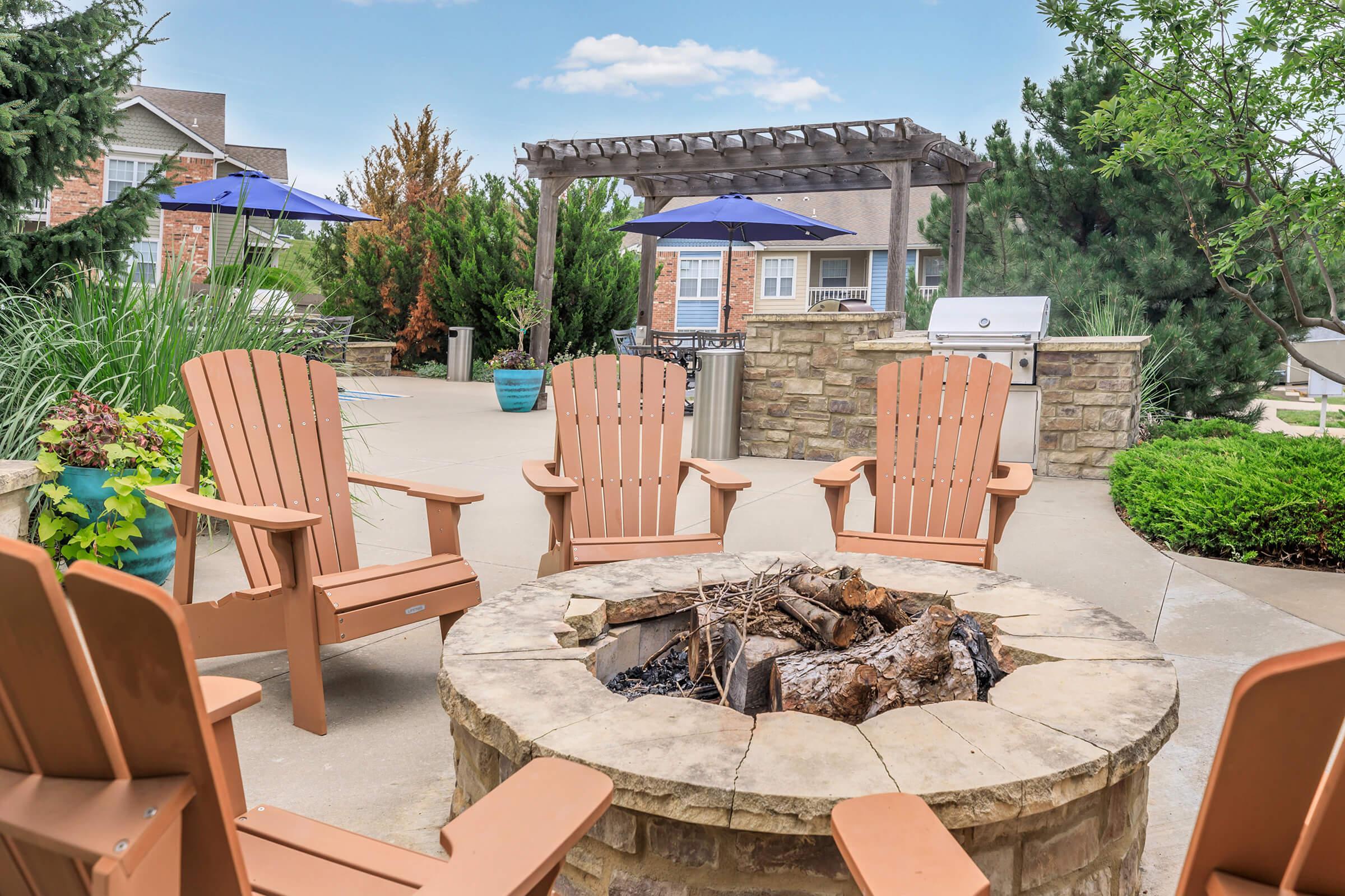 A cozy outdoor area featuring a stone fire pit surrounded by four wooden Adirondack chairs. There are blue umbrellas providing shade, lush greenery in the background, and a glimpse of a grill and a patio area, creating an inviting atmosphere for relaxation and gatherings.