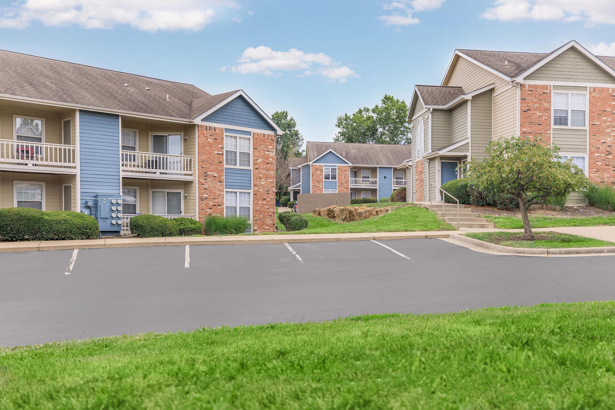 Two multi-unit residential buildings in a suburban area. One building features blue siding with balconies, while the other is brick and light green. A well-maintained lawn with a gentle slope and small bushes is in the foreground. The sky is partly cloudy, creating a peaceful atmosphere.