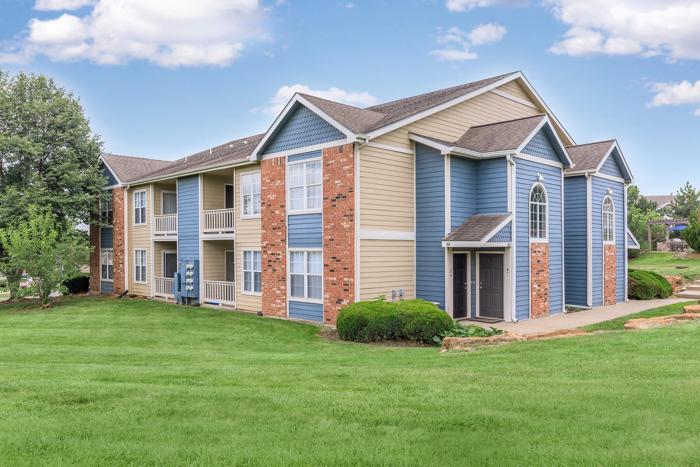 A modern two-story apartment building featuring a mix of blue siding and brick accents. The property is surrounded by well-maintained green grass and shrubs, with a clear blue sky above and fluffy clouds. The building has multiple balconies and windows, showcasing a clean and inviting residential environment.