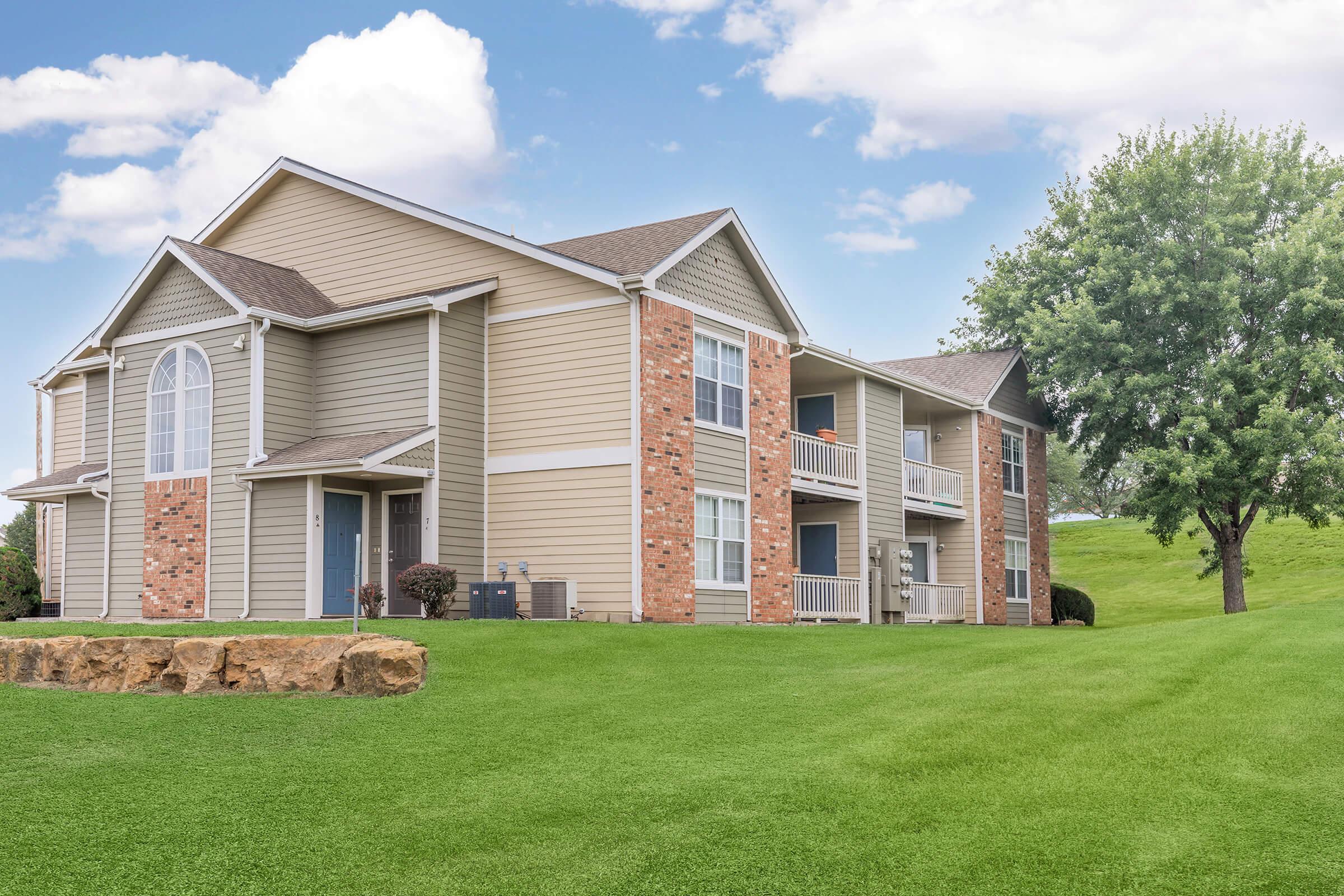 A two-story residential building with a mix of light-colored siding and stone accents, surrounded by green grass and a tree. The building features balconies, large windows, and a well-maintained lawn. The sky is partly cloudy, creating a bright and inviting atmosphere.