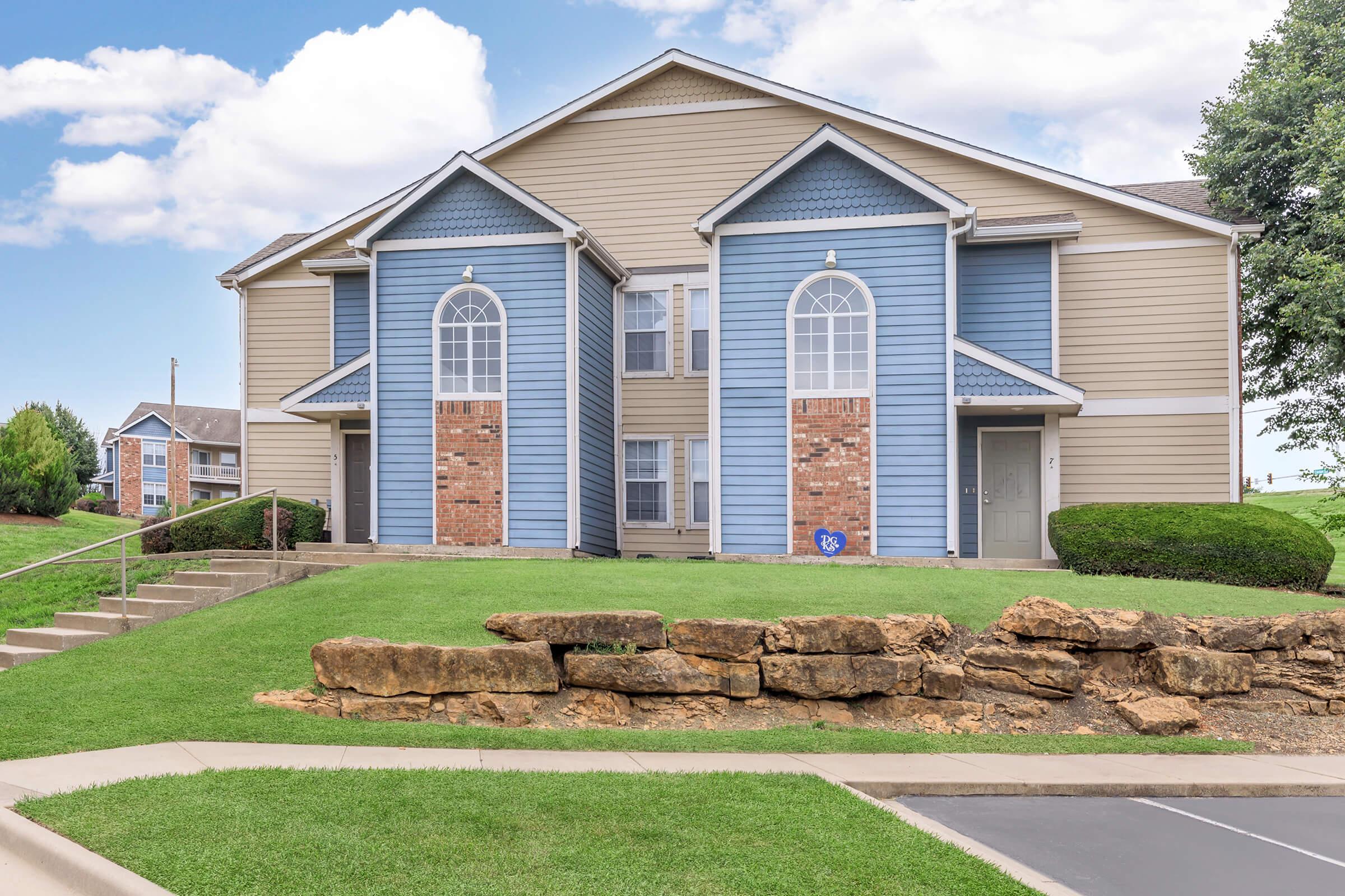 Two-story apartment building with blue and beige siding, featuring decorative brick accents. There is a neatly landscaped area in front with a stone wall and grassy lawn. The sky is partly cloudy, and additional buildings are visible in the background.