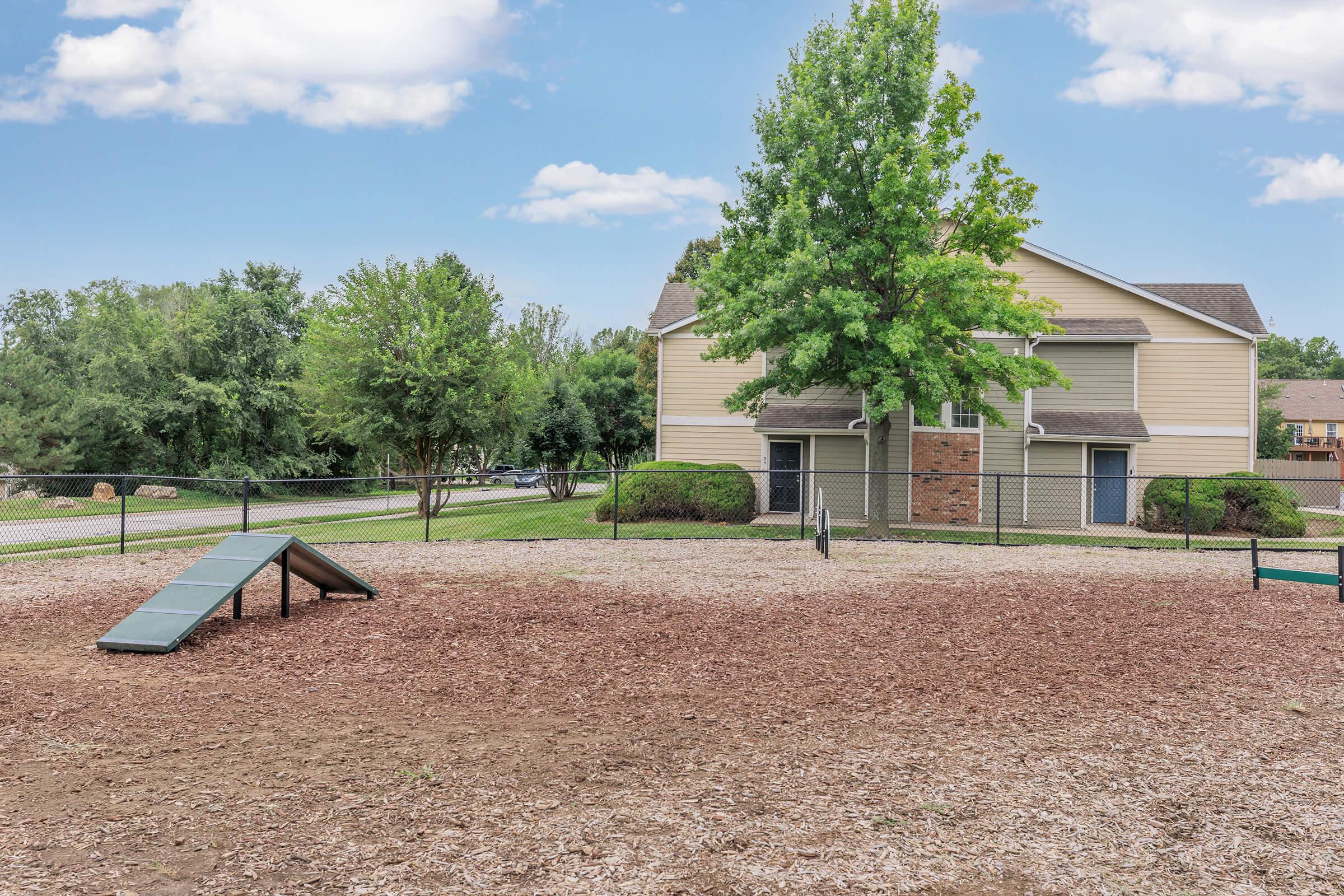 A fenced dog park featuring a small agility ramp, surrounded by wood chip flooring. In the background, there is a two-story residential building and trees, with a grassy area. The sky is clear with a few clouds overhead.