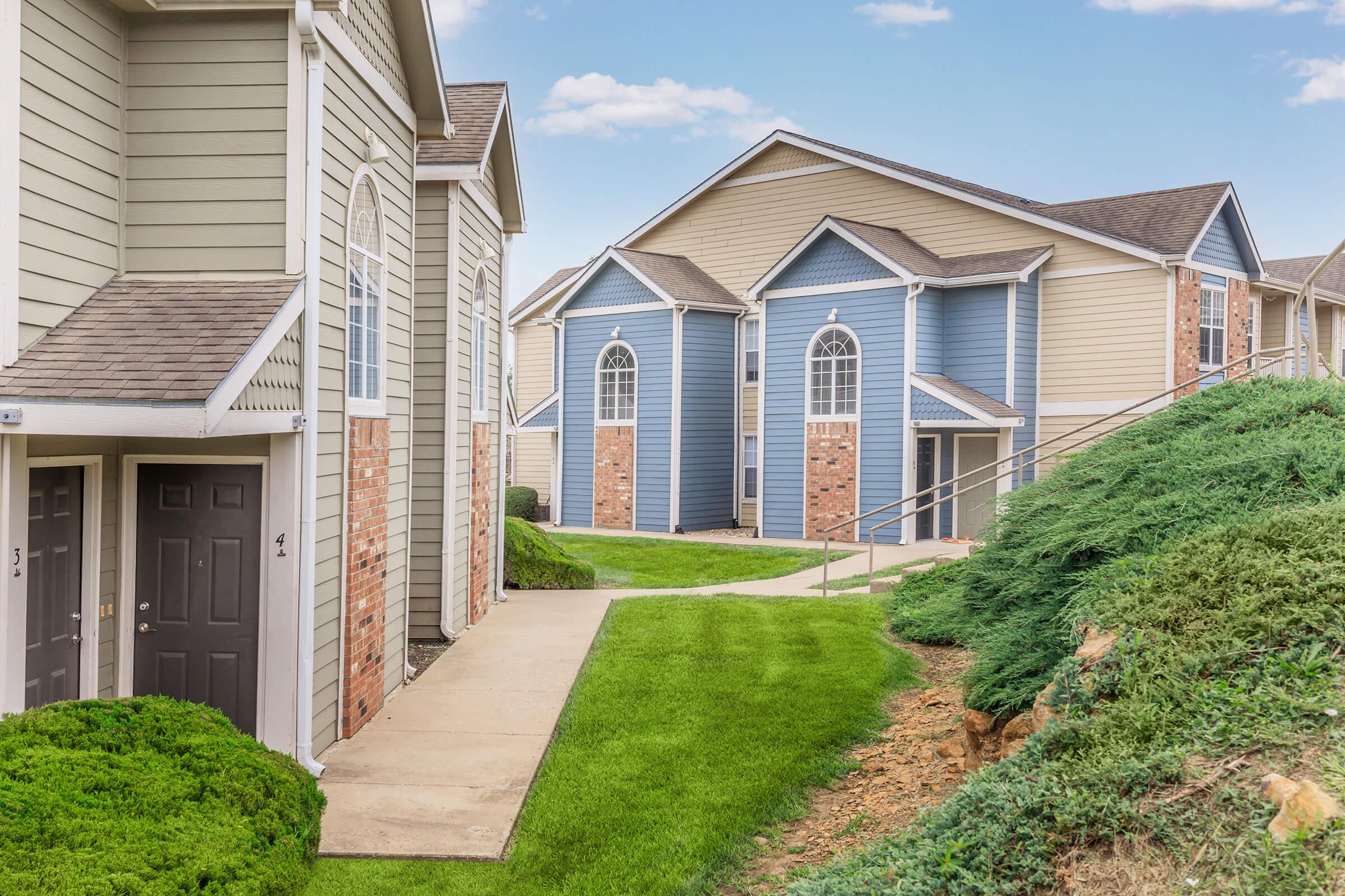 Two multi-unit residential buildings with varying exterior colors, including beige and blue, are situated along a landscaped pathway. There is manicured greenery and a gentle slope in the foreground, with clear skies visible in the background.