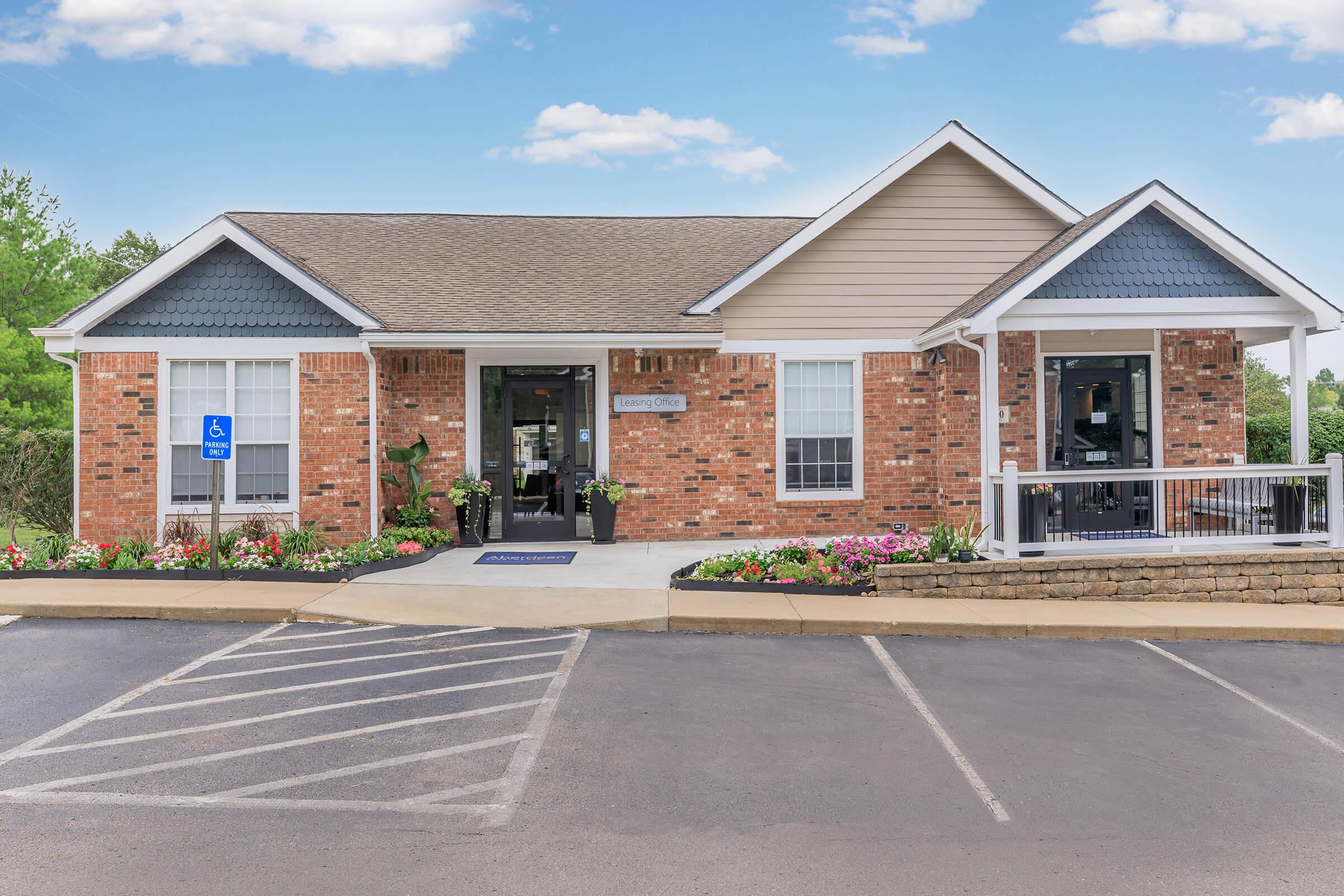 A brick building with a sloped roof featuring a welcoming entrance, surrounded by vibrant flower beds and greenery. The parking area includes designated spaces, and there are ramps for accessibility, creating an inviting atmosphere. Clear blue skies add to the overall pleasant scene.