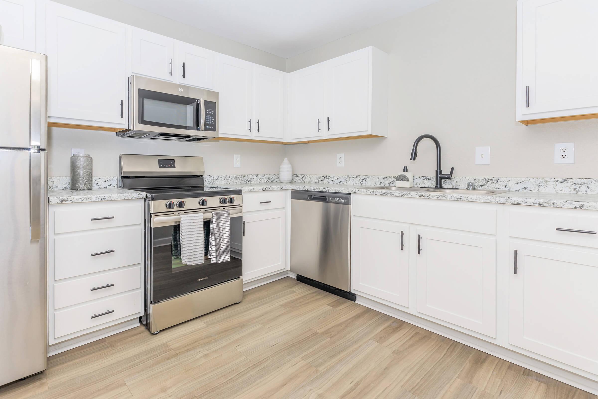Modern kitchen featuring stainless steel appliances, including an oven, microwave, and dishwasher. The white cabinets complement the light gray walls and a granite countertop. The space has a clean look with wooden flooring and decorative items on the countertop.