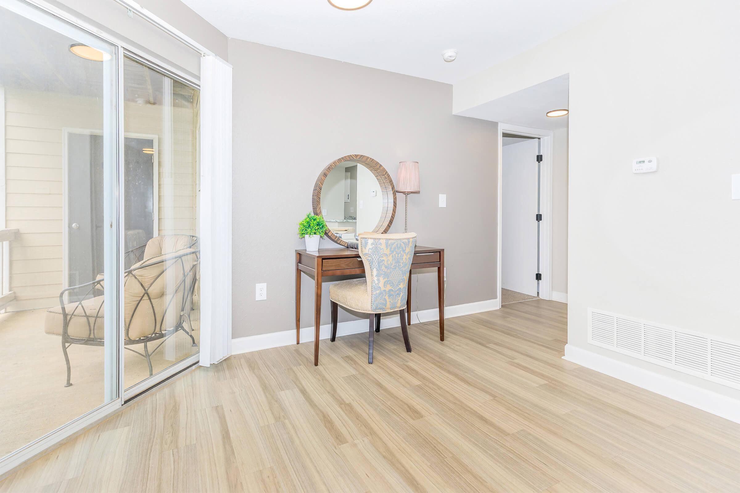 Bright, modern living space featuring a wooden floor, a small desk with a decorative chair, and a round mirror on the wall. Natural light enters through sliding glass doors leading to a balcony. Minimalist décor and light-colored walls create an inviting atmosphere.