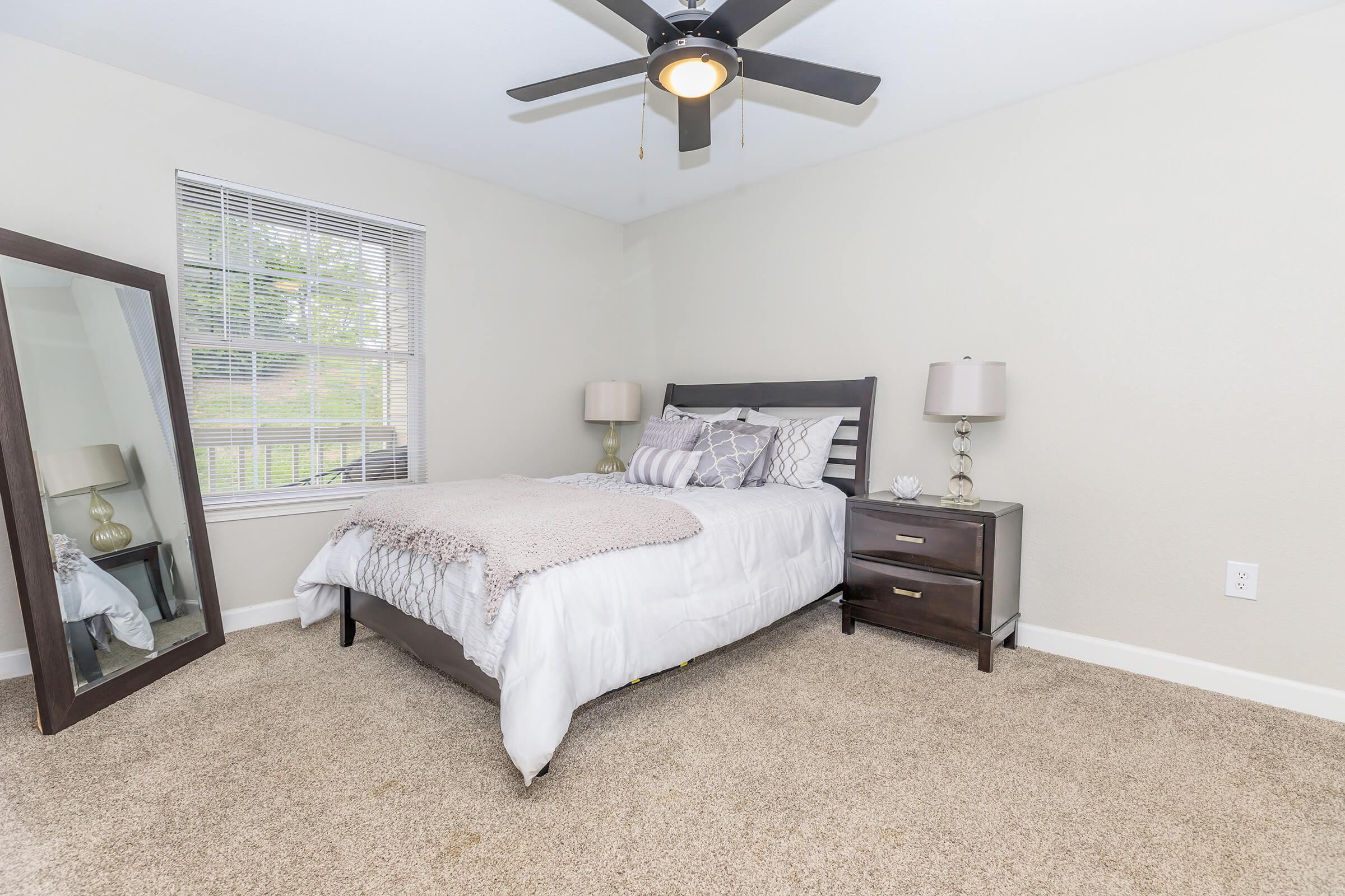 A cozy bedroom featuring a bed with white and gray bedding, two nightstands with lamps, a large mirror reflecting the room, soft beige carpet, and a ceiling fan. Natural light comes through the window, highlighting the neutral-colored walls and inviting atmosphere.