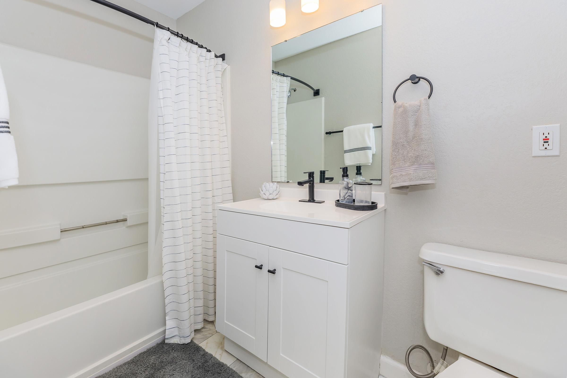 A modern bathroom featuring a white bathtub with a shower curtain, a sleek white vanity with a sink and mirror, a towel hanging on a rack, and a toilet. The walls are painted in a light color, and a gray rug is placed on the floor. Natural light is coming from an overhead fixture.
