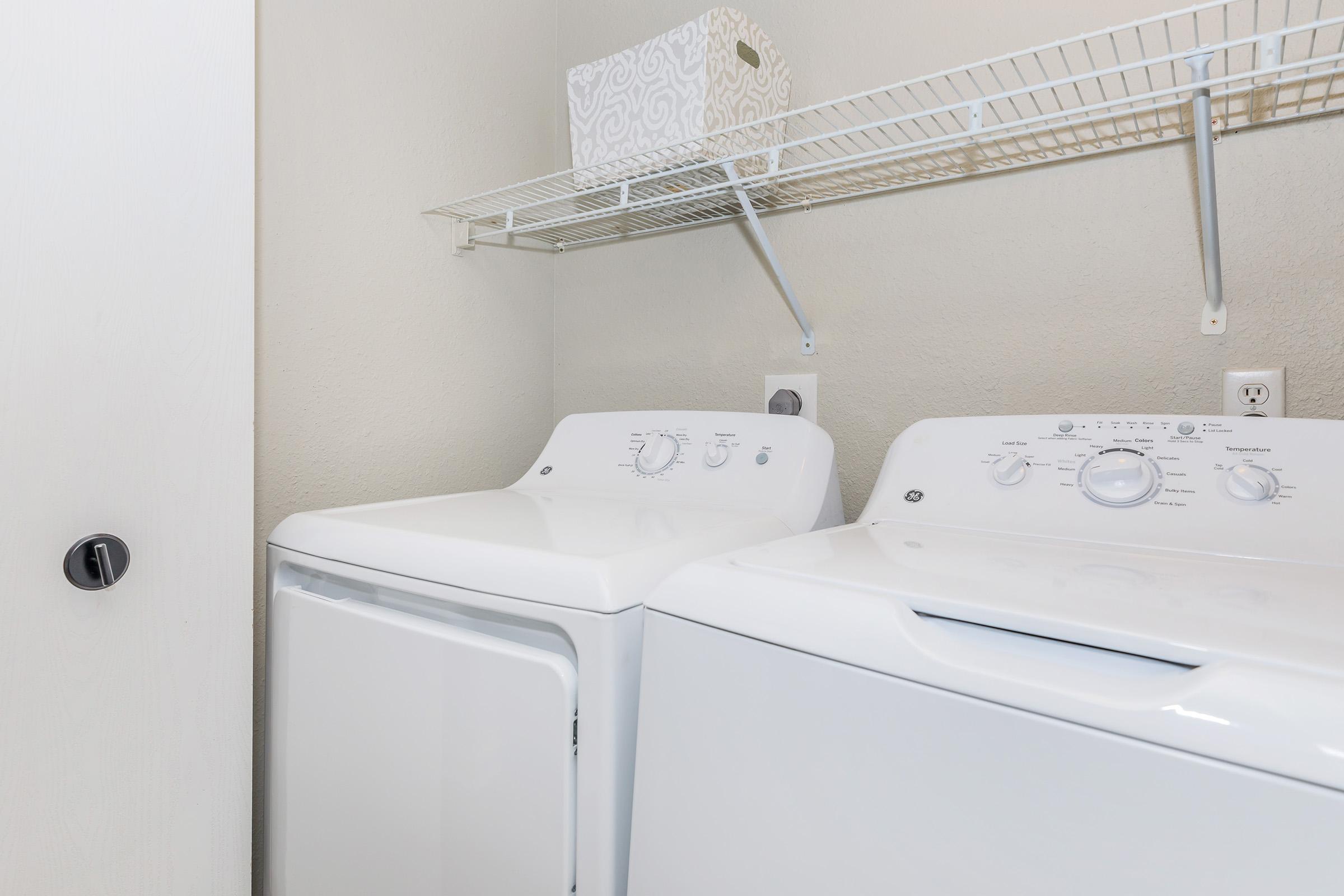 A laundry room featuring a stacked washer and dryer in white. Above the appliances, there is a metal wire shelf with a decorative storage basket. The walls are a neutral color, and a door handle is visible on the left side of the image. The overall space is clean and organized.