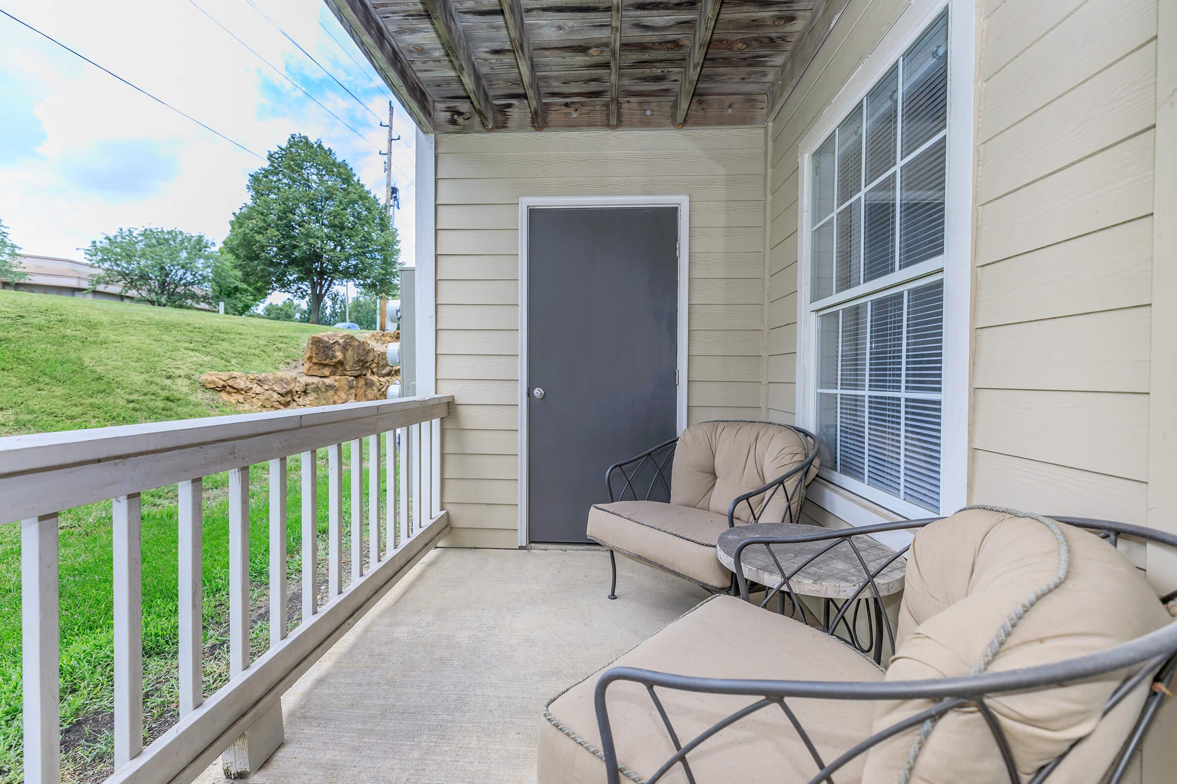 A small outdoor balcony featuring two cushioned chairs and a table, with a view of green grass and trees in the background. A closed gray door is visible, along with a wooden railing. The sky is partly cloudy, adding a bright atmosphere to the space.