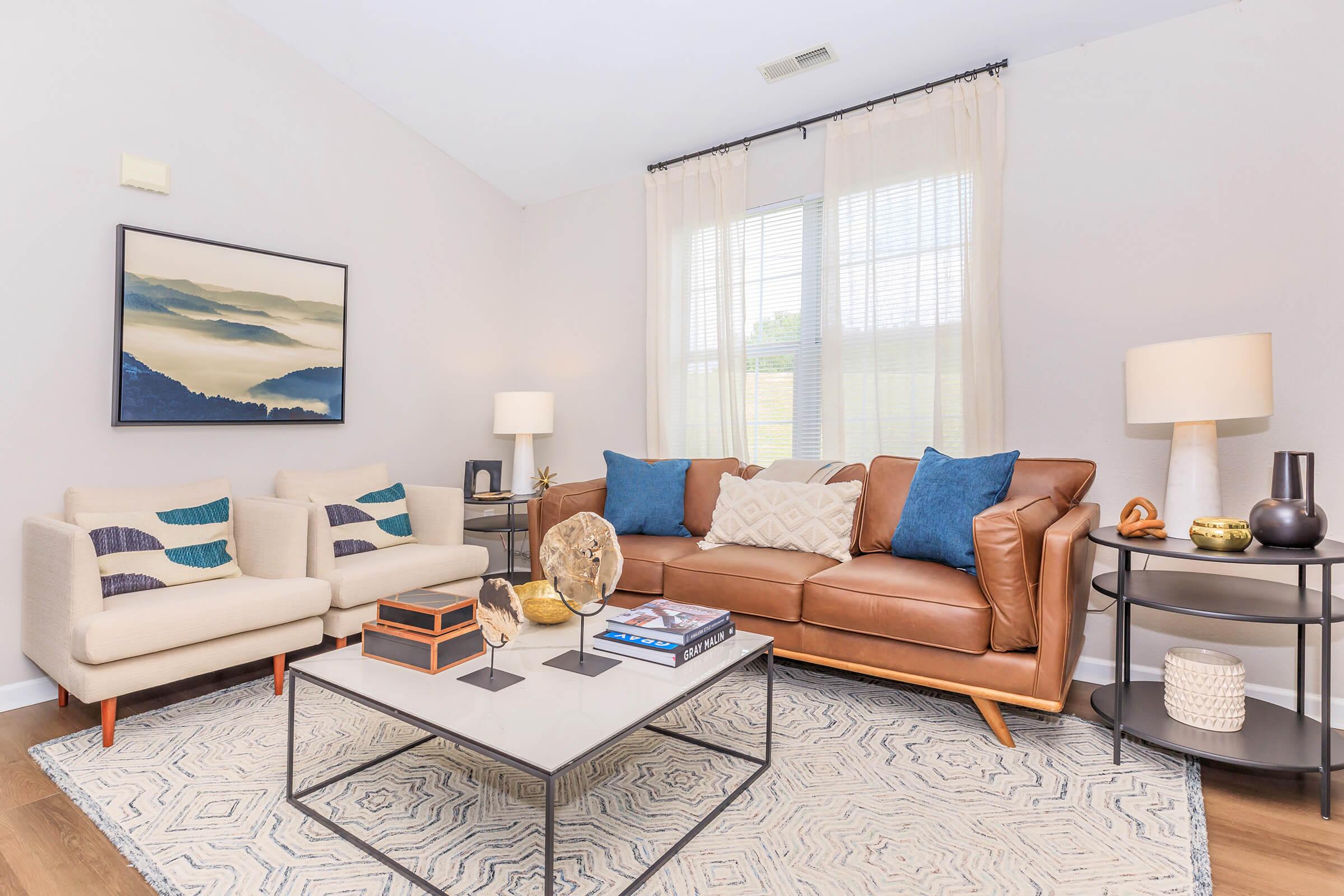 A stylish living room featuring a brown leather sofa and two light-colored armchairs with decorative cushions. A coffee table with decor items sits in the center, and there are elegant curtains framing a window. A framed landscape artwork hangs on the wall, adding a modern touch to the space.