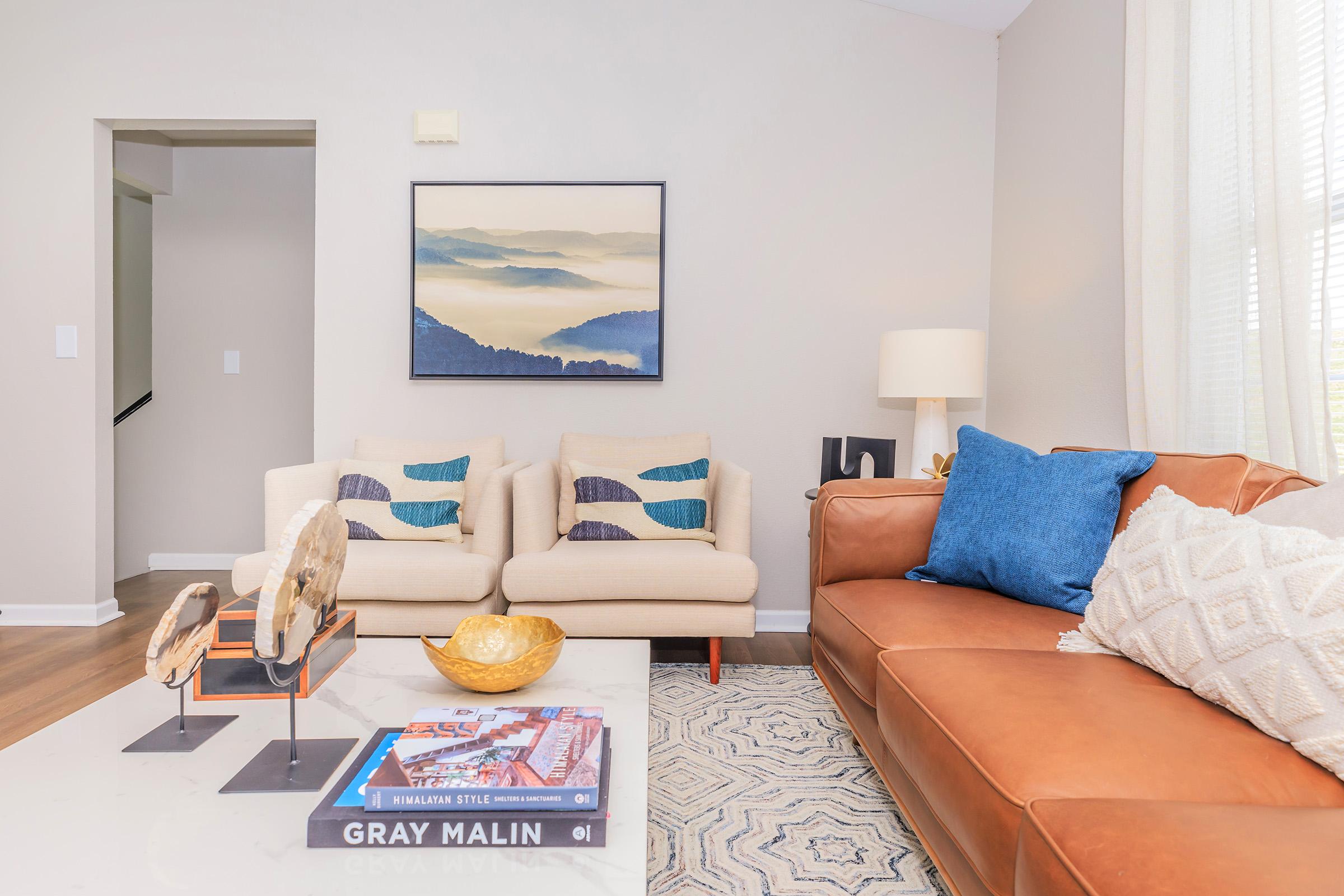 A stylish living room featuring two cream-colored chairs with blue and turquoise cushions, a brown leather sofa with a blue throw pillow, and a coffee table displaying books and a decorative bowl. A landscape painting hangs above the sofa, and natural light filters through sheer curtains.