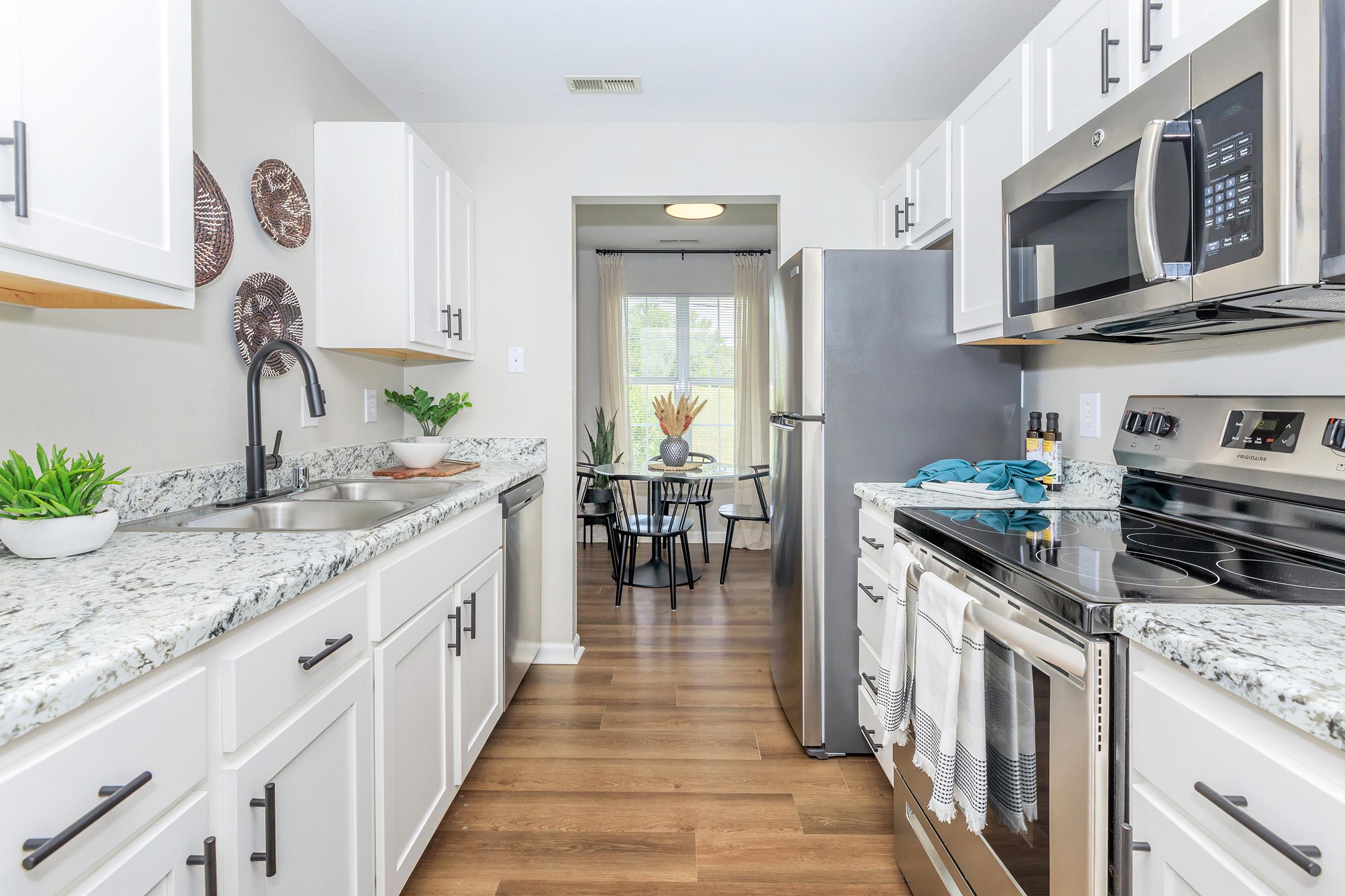 Bright modern kitchen with white cabinetry and granite countertops. Features stainless steel appliances, a sink with a black faucet, and decorative plates on the wall. Opens to a dining area with a table and chairs, allowing natural light through a large window.
