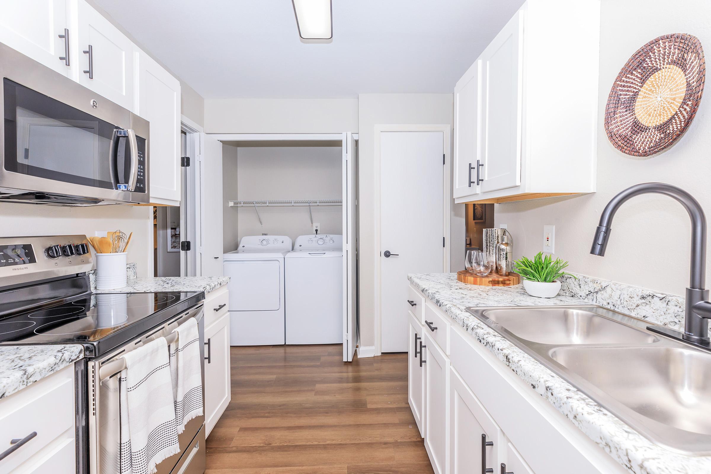 A modern kitchen featuring white cabinets, stainless steel appliances, and a granite countertop. An adjoining laundry area is visible, equipped with a washer and dryer. The space is well-lit with a neutral color palette and a touch of greenery, creating a clean and inviting atmosphere.