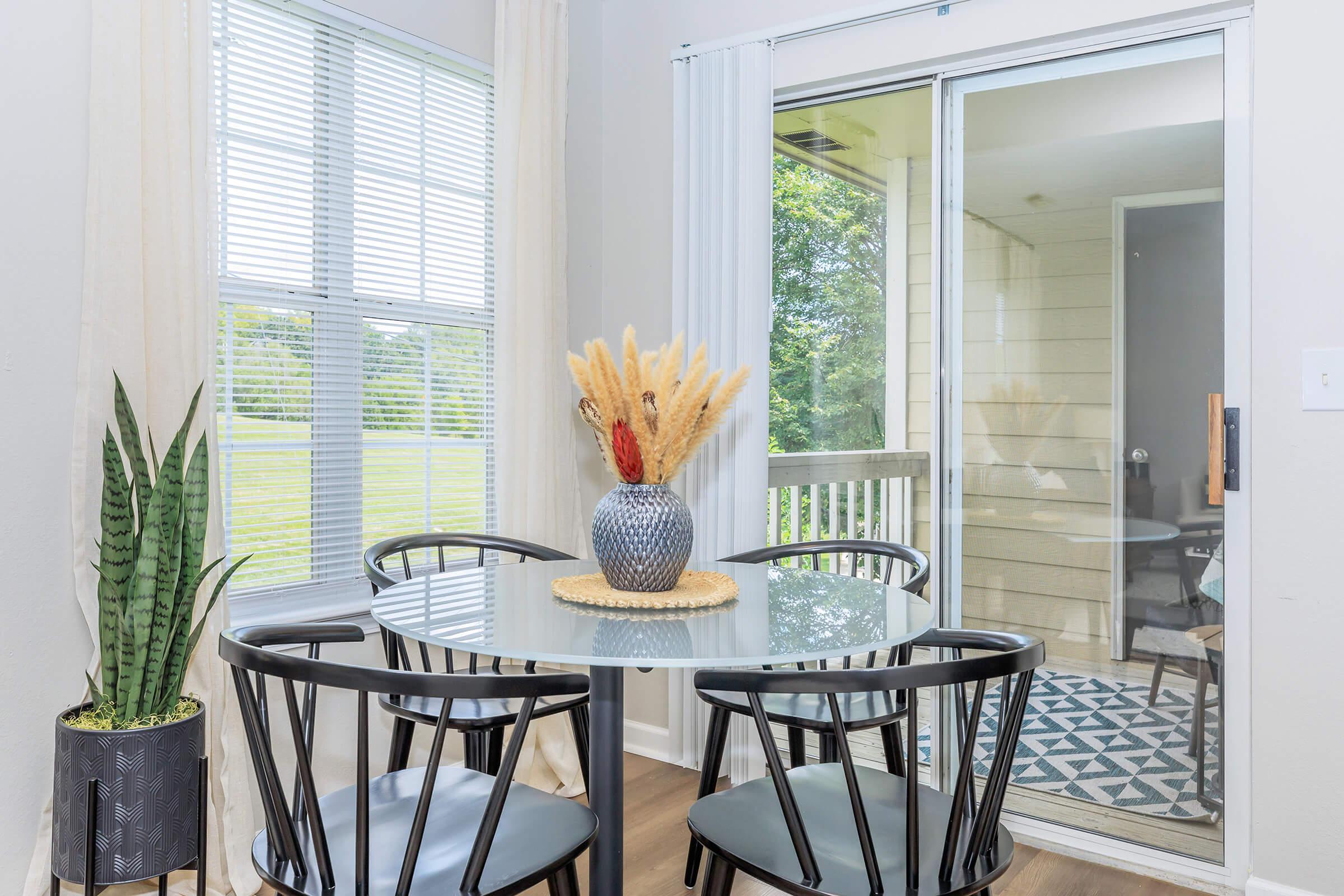 A bright dining area featuring a round glass table with four black chairs. A vase with dried flowers sits on the table. There’s a tall green plant in a pot next to the window, which has sheer curtains. A sliding glass door leads to an outdoor view of green space.