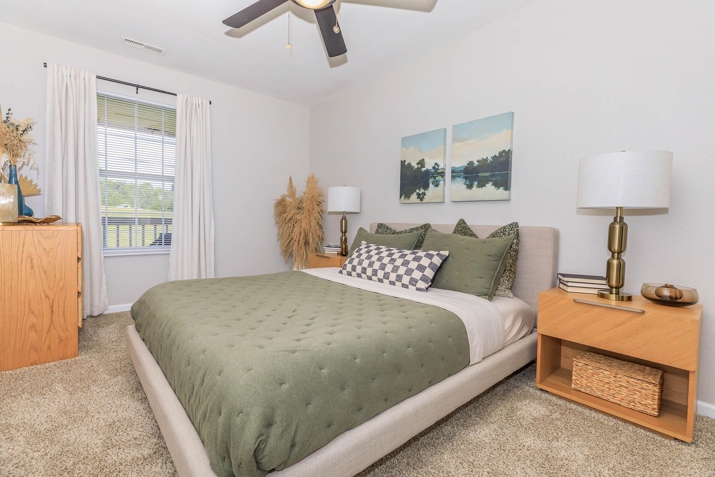A cozy bedroom featuring a queen-sized bed with a green quilt and patterned pillows. There are wooden nightstands on either side, each with a lamp. A plant decorates the room, and large windows let in natural light, complemented by neutral-colored walls and carpet. Artwork above the bed adds a touch of color.