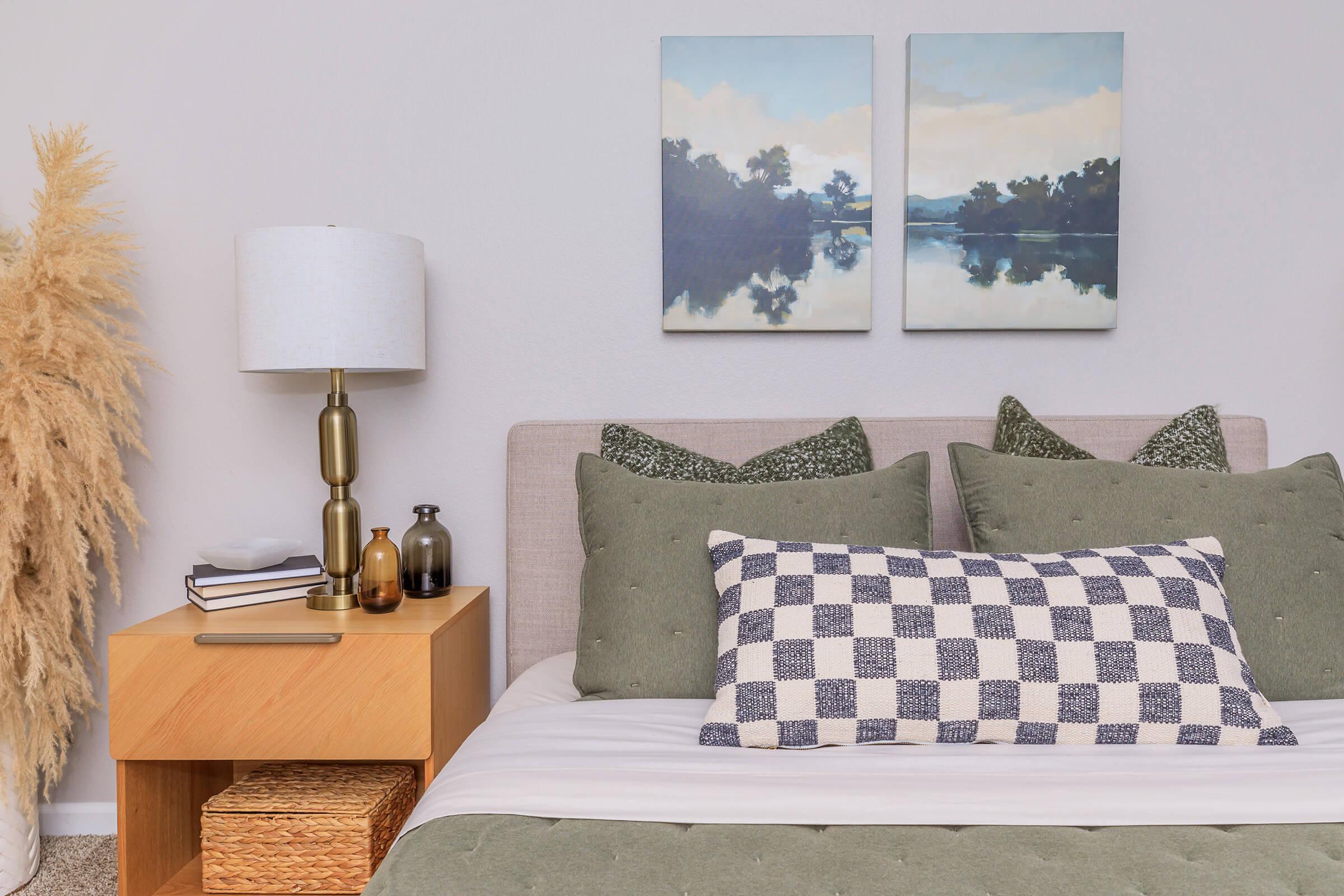 A cozy bedroom scene featuring a neatly made bed with green and patterned pillows. Adjacent is a wooden nightstand with a lamp, decorative bottles, and stacked books. On the wall, two landscape paintings depict a serene lakeside view, complemented by a decorative plant in the corner.
