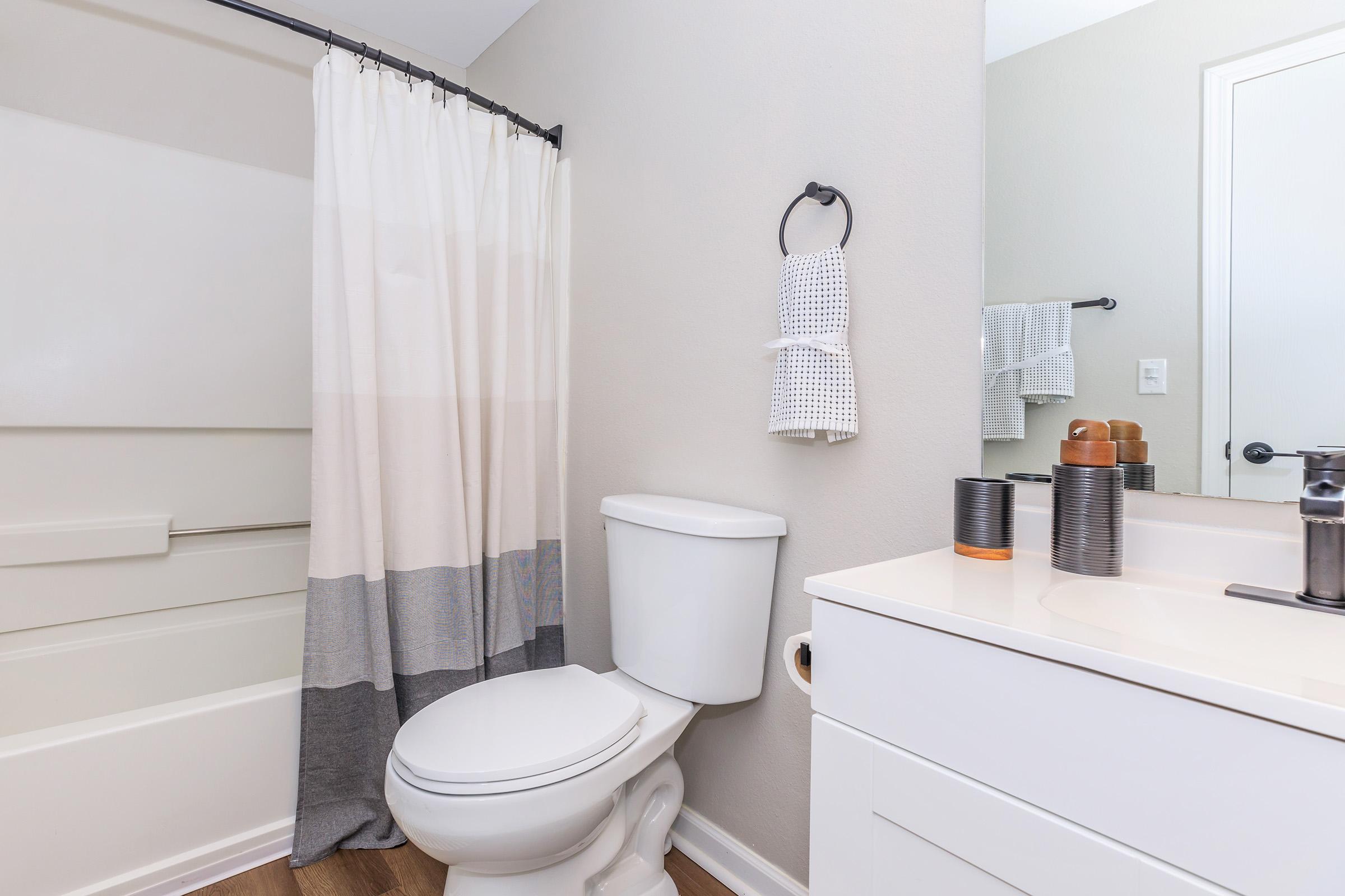A clean and modern bathroom featuring a shower with a curtain, a white toilet, and a vanity with a sink. There are neatly arranged towels and bathroom accessories on the wall and countertop, with neutral-colored walls and wooden flooring creating a fresh and inviting atmosphere.
