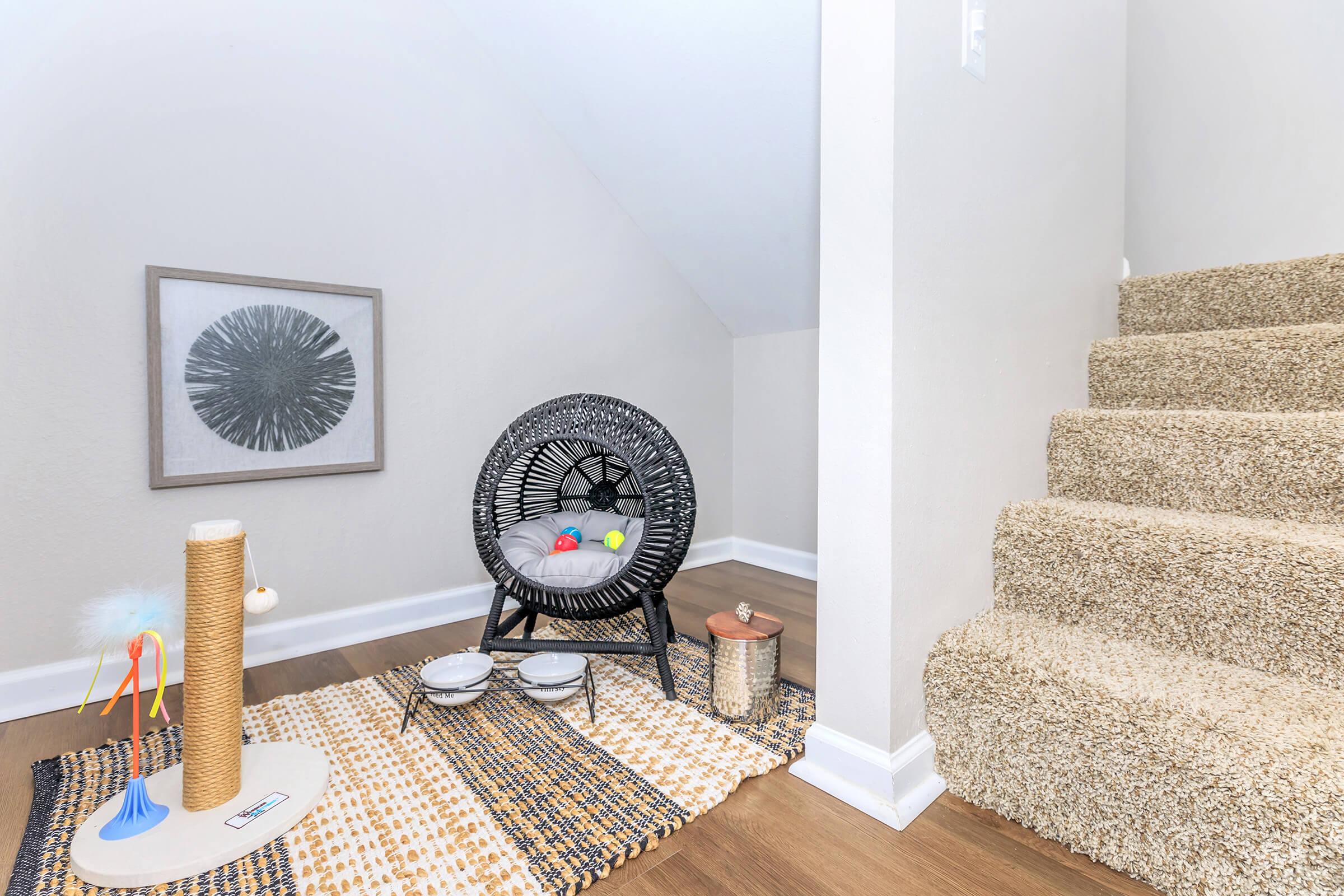 Cozy corner of a home featuring a black wicker chair with cushions, a small rug, and a cat scratching post. A few colorful toys are scattered around. Stairs are visible in the background, leading to an upper level, and a framed decorative wall art piece hangs nearby.