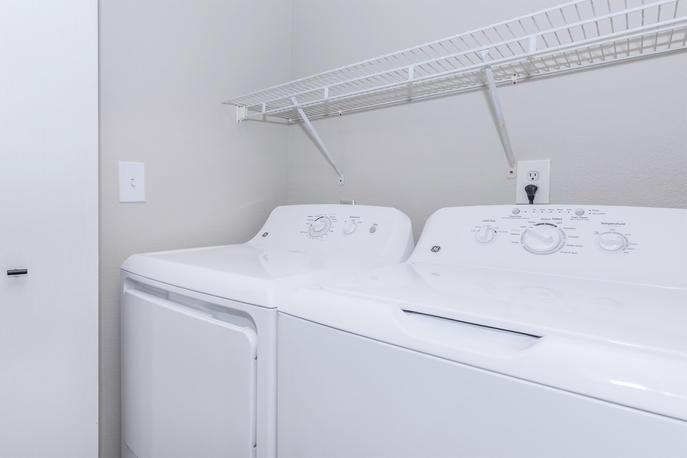 A clean laundry room featuring a white washer and dryer side by side, with a wall-mounted shelf above them. The walls are painted in a light color, and there is a door partially visible on the left. The appliances have control dials and settings clearly displayed.