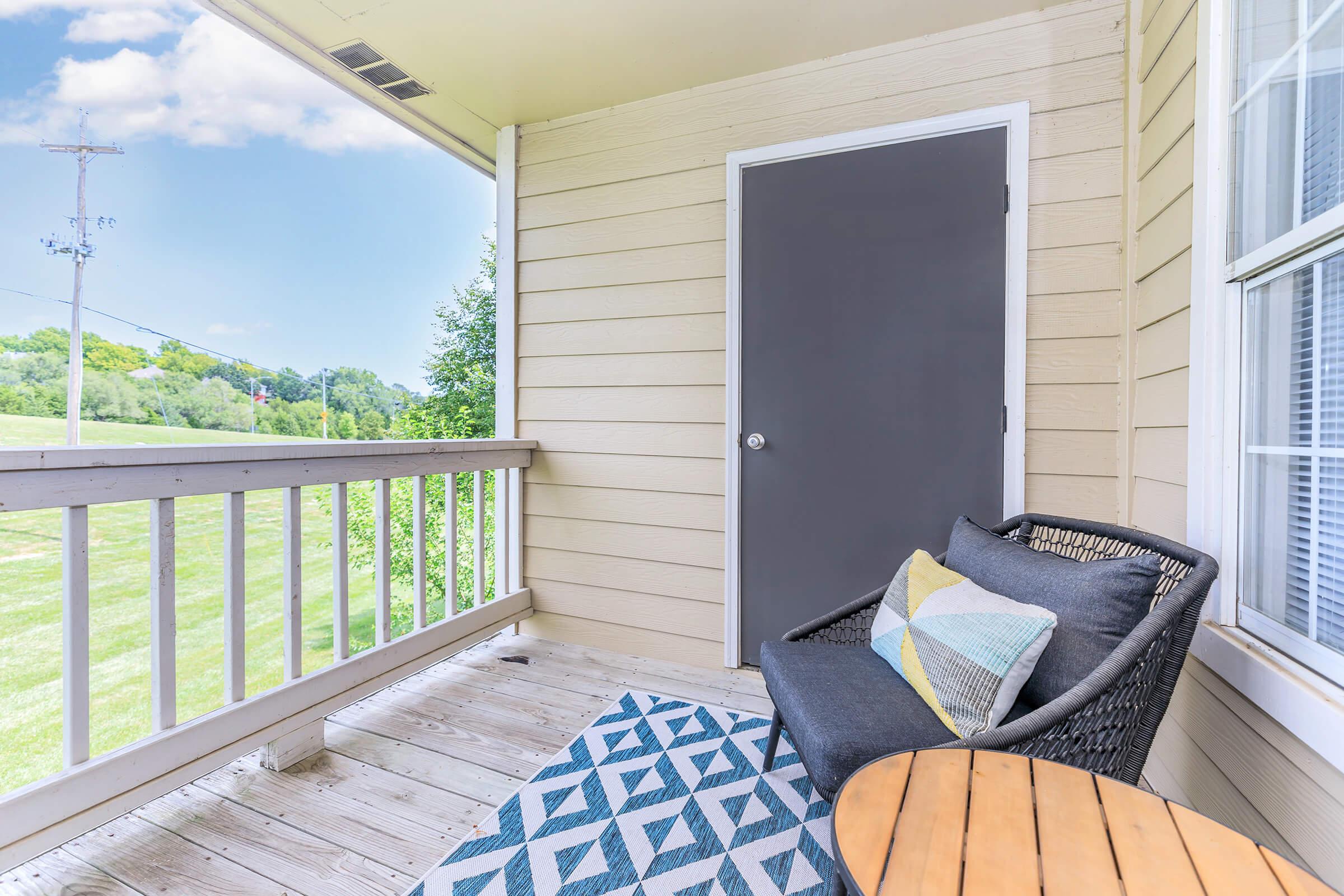 A cozy balcony featuring a gray door, a comfortable wicker chair with a colorful pillow, and a round wooden table. The floor is adorned with a patterned rug, and there are lush green fields visible in the background under a bright blue sky.