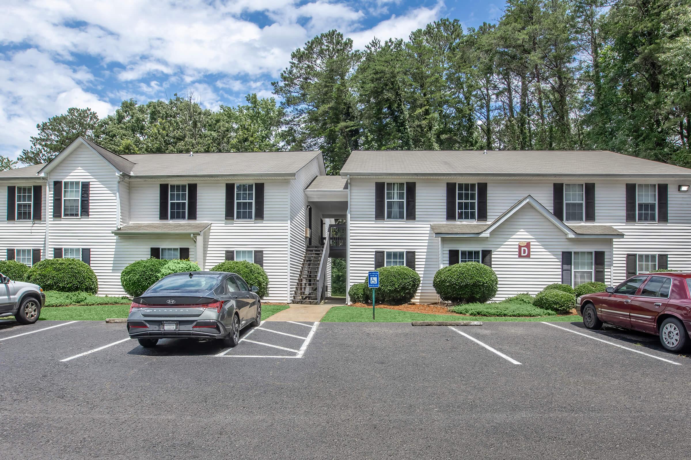 A two-story apartment building with white siding set among green trees. The front features several parking spaces, including accessible parking. The building has multiple windows and a central entrance, with well-maintained landscaping. Bright blue sky with some clouds is visible above.