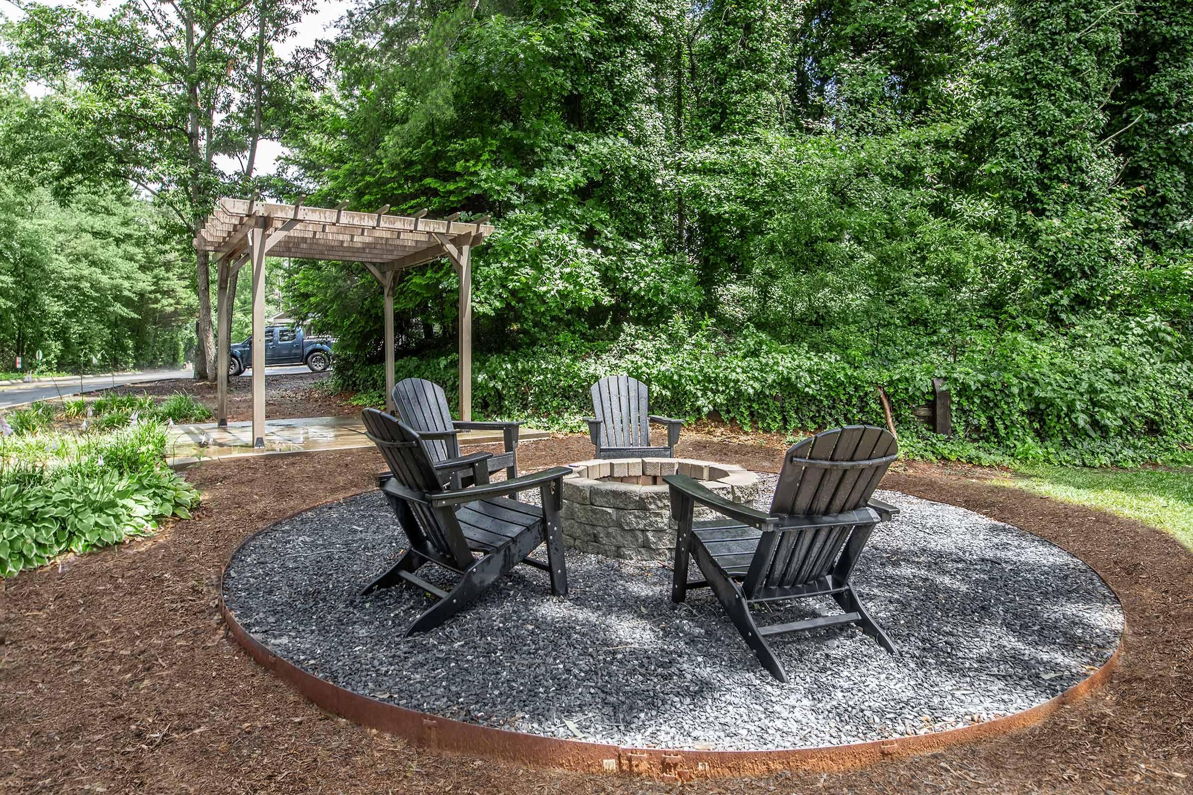 A circular fire pit area featuring four black Adirondack chairs arranged around a stone fire pit, surrounded by gravel and mulch. Lush greenery and trees provide a natural backdrop, with a wooden pergola partially visible nearby. A vehicle can be seen in the background along a road.