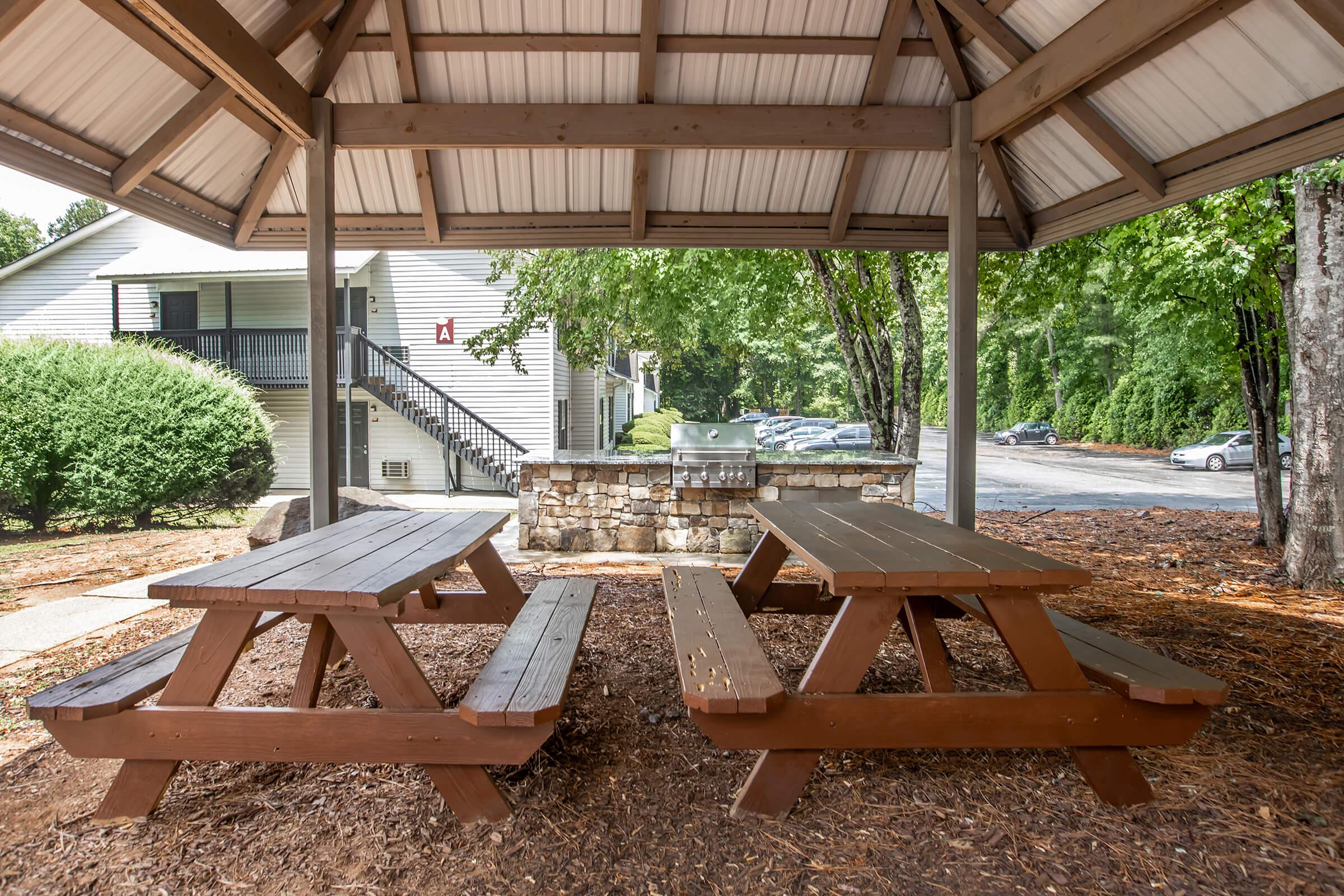 A covered picnic area featuring multiple wooden picnic tables surrounded by mulch, with green trees in the background. In the distance, a walkway leads to a multi-unit building and a parking area.