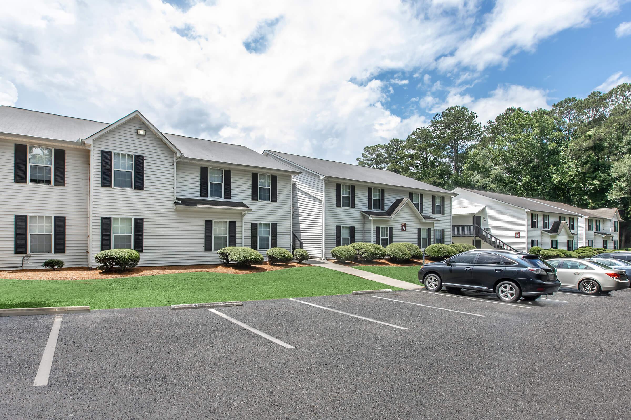 A row of two-story apartment buildings with light-colored siding and black shutters. In the foreground, there is a parking lot with several parked cars. The landscape features trimmed shrubs and green grass, under a partly cloudy sky.
