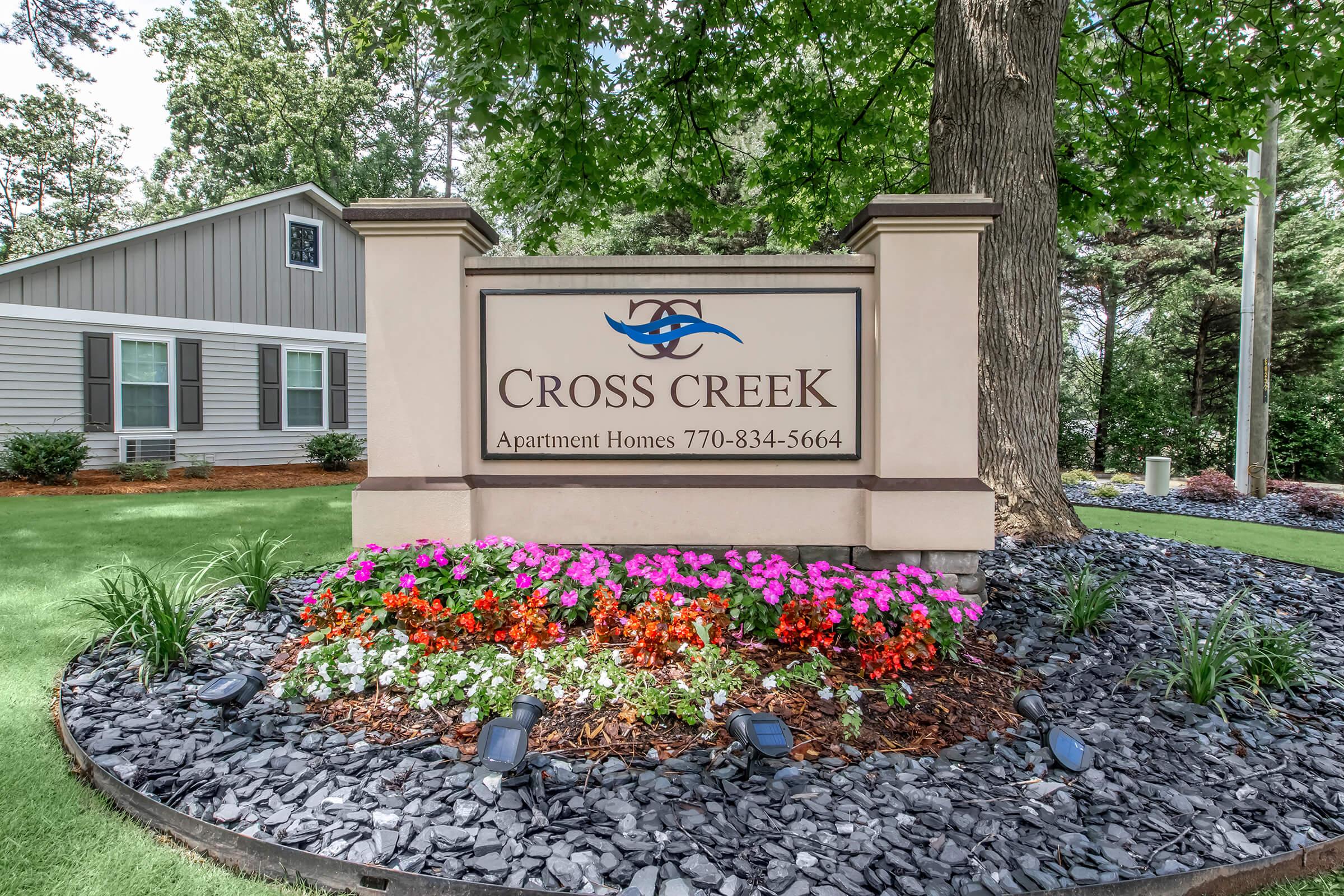 Sign for Cross Creek Apartment Homes with a decorative flower bed. The sign features the name "CROSS CREEK" prominently, along with a logo and contact number (770-834-5664). Surrounding the sign are colorful flowers and black stones, set against a backdrop of green grass and trees.