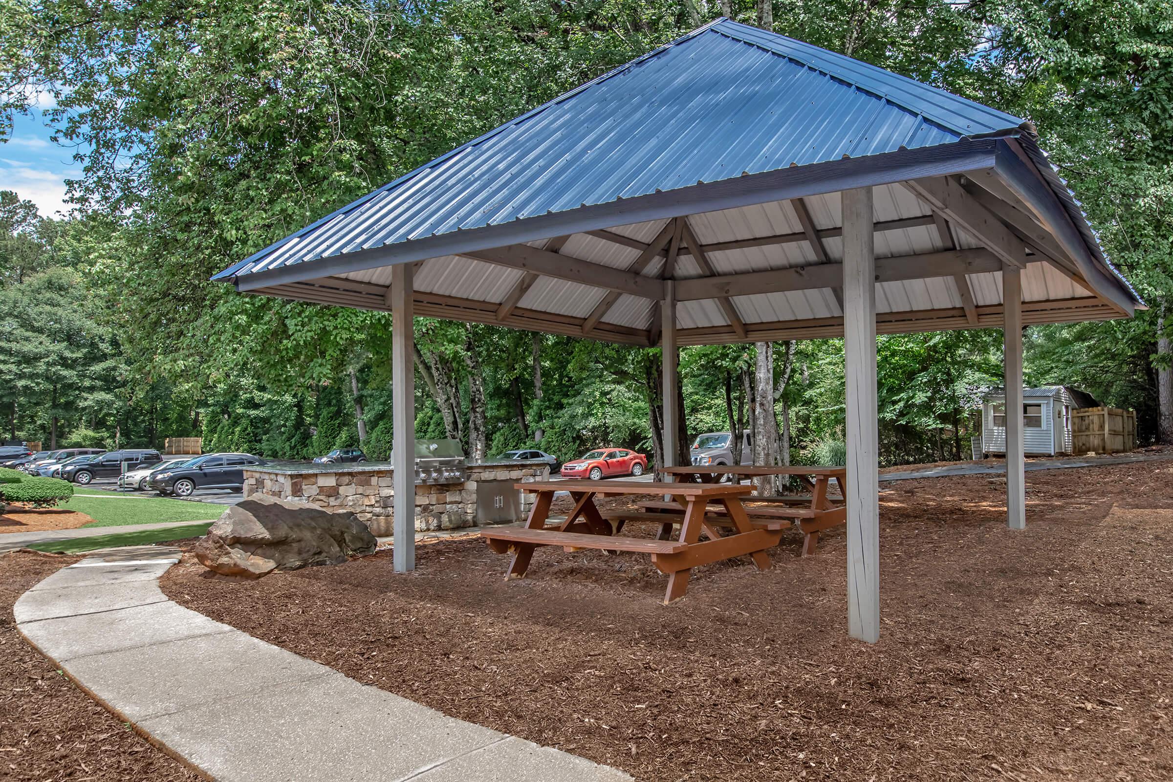 A covered picnic area with a blue roof is situated in a wooded park. Underneath, there are wooden picnic tables. A concrete path leads to the area, with parked cars visible in the background and landscaped greenery surrounding the space.