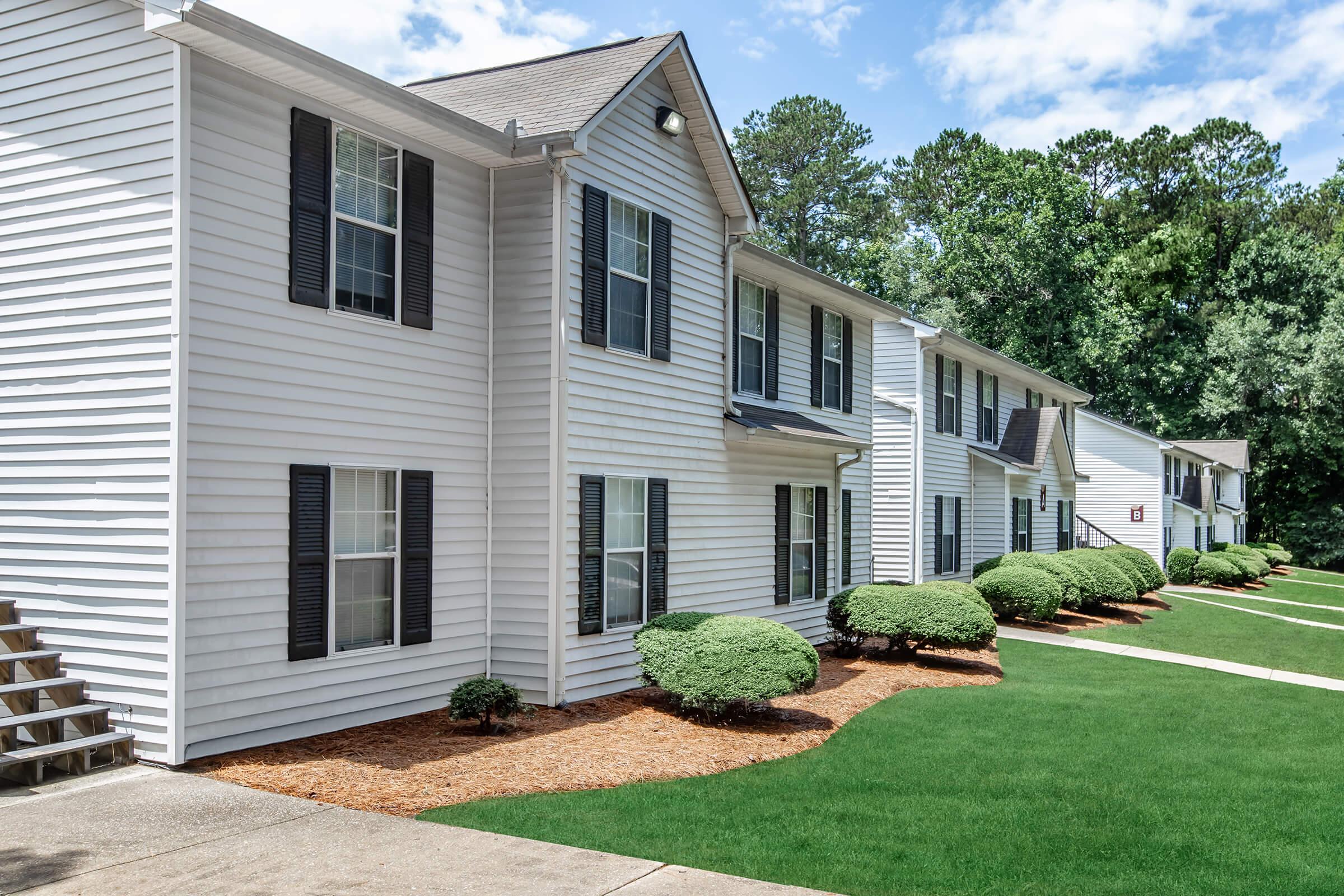 A row of multi-unit residential buildings with white siding and dark shutters. The landscaping features neatly trimmed bushes and green grass. The blue sky with fluffy clouds is visible in the background, adding a bright and inviting atmosphere to the scene.