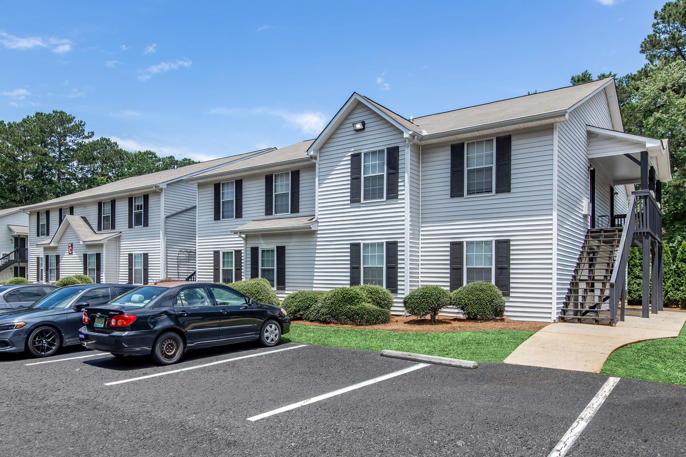Two-story residential apartment building with white siding and black shutters. A small staircase is visible on the right side. In the foreground, there are parked cars and neatly trimmed shrubs. The area has green grass and a clear blue sky.