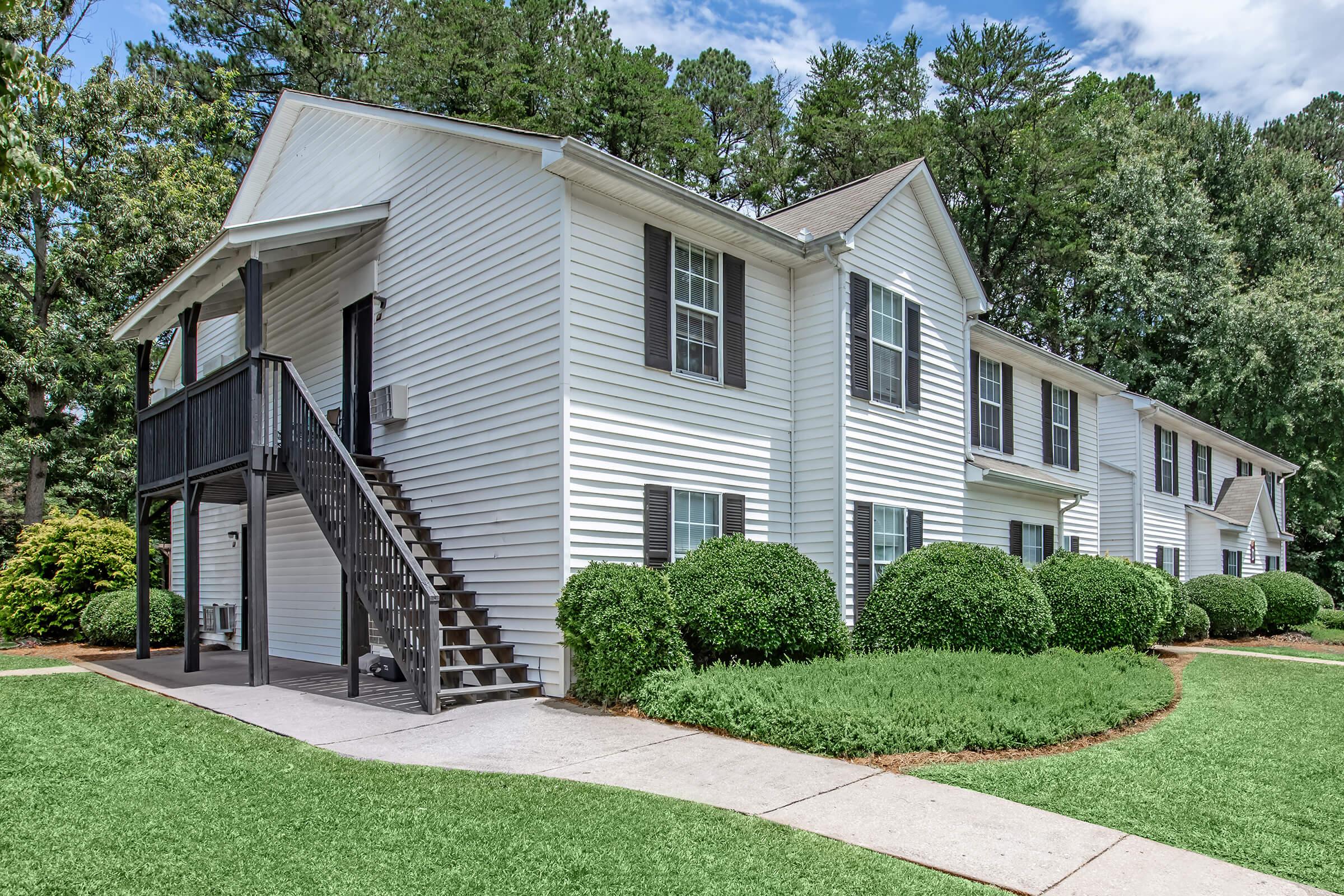 View of a two-story apartment building featuring white siding and black shutters. The building is flanked by neatly trimmed bushes and grassy areas. A staircase leads to the second floor, and the sky is partly cloudy, suggesting a pleasant day.