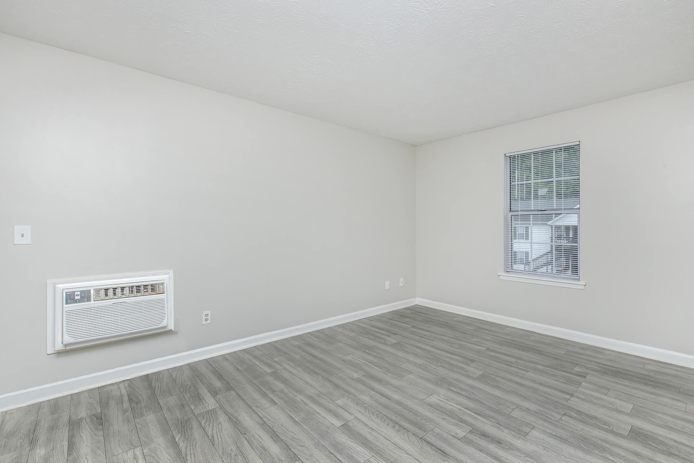 A spacious, empty room with light gray walls and a window. There is an air conditioning unit on the left wall, and the floor features wood-like tiles. Natural light enters through the window, providing a bright atmosphere in the uncluttered space.