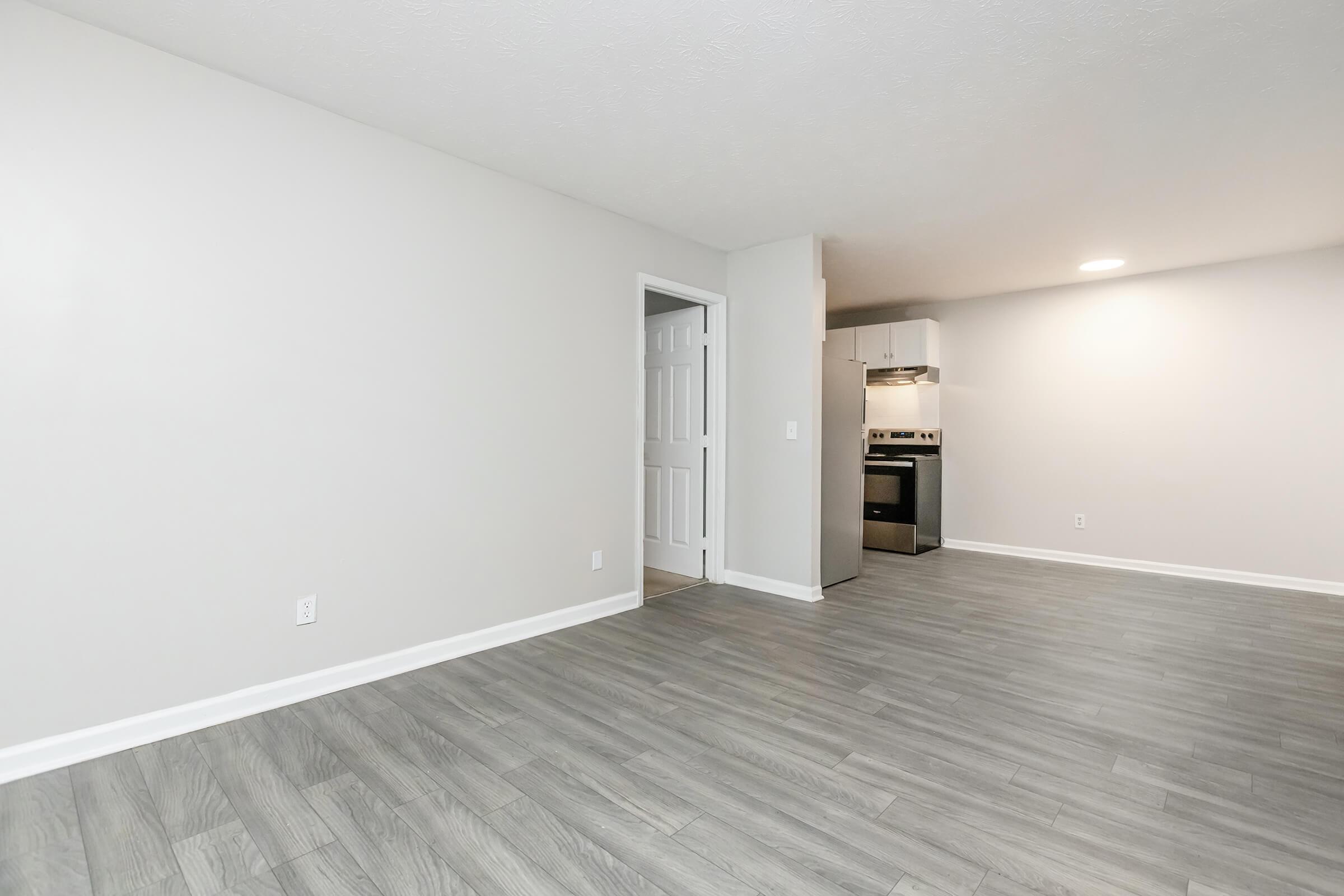 A spacious, light-colored room with gray laminate flooring and a neutral wall. A doorway on the left leads to another room, while the right side features a kitchen area with appliances visible, including a stove and refrigerator. The overall aesthetic is modern and minimalistic.