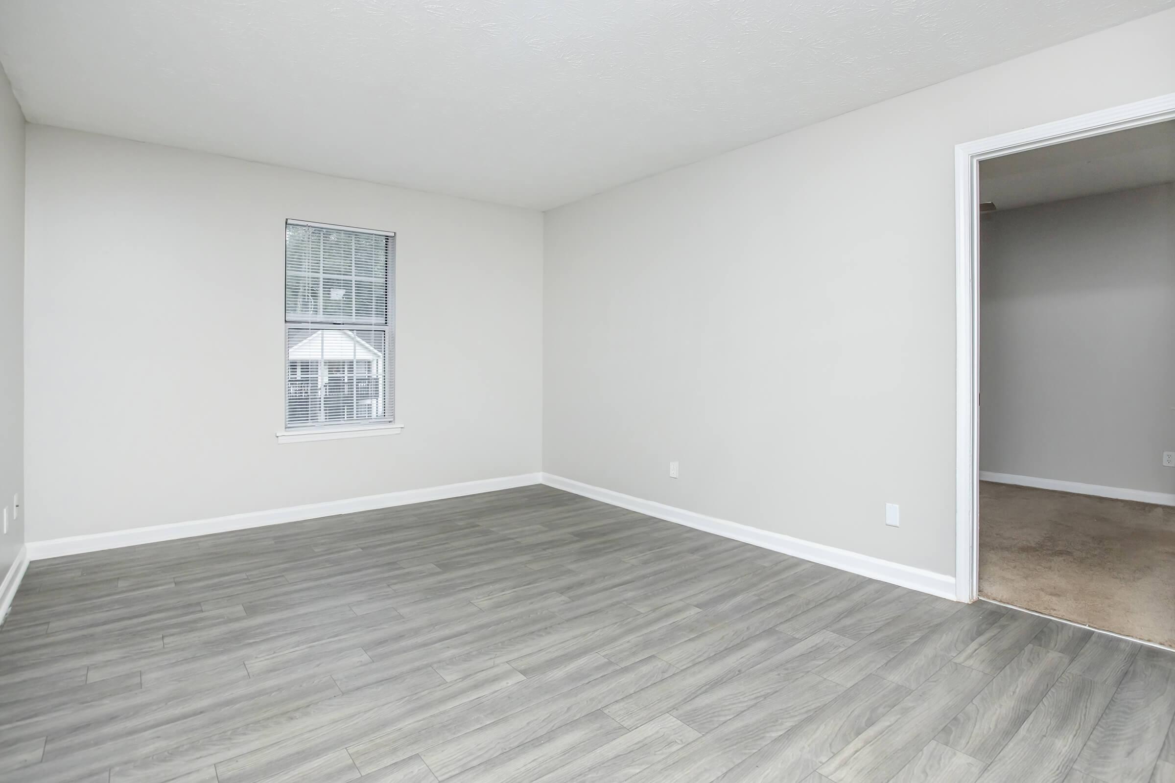 A spacious, empty room with light gray walls and hardwood-style flooring. A window with blinds lets in natural light, and a doorway leads to another room. The overall ambiance is bright and airy, making it suitable for various decorating styles.