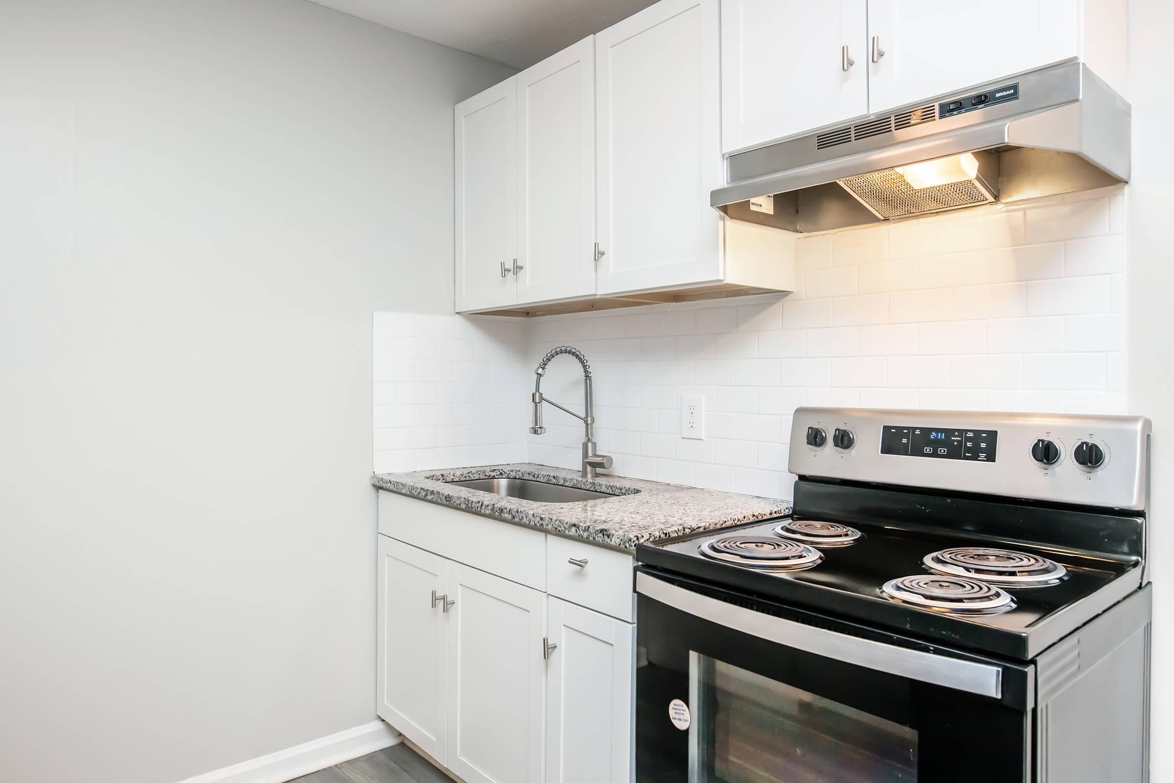 A modern kitchen featuring white cabinets, a granite countertop, a stainless steel sink with a pull-down faucet, and a black stove with four burners. The backsplash is white subway tile, and there is an overhead range hood. The walls are painted light gray, creating a clean and contemporary look.