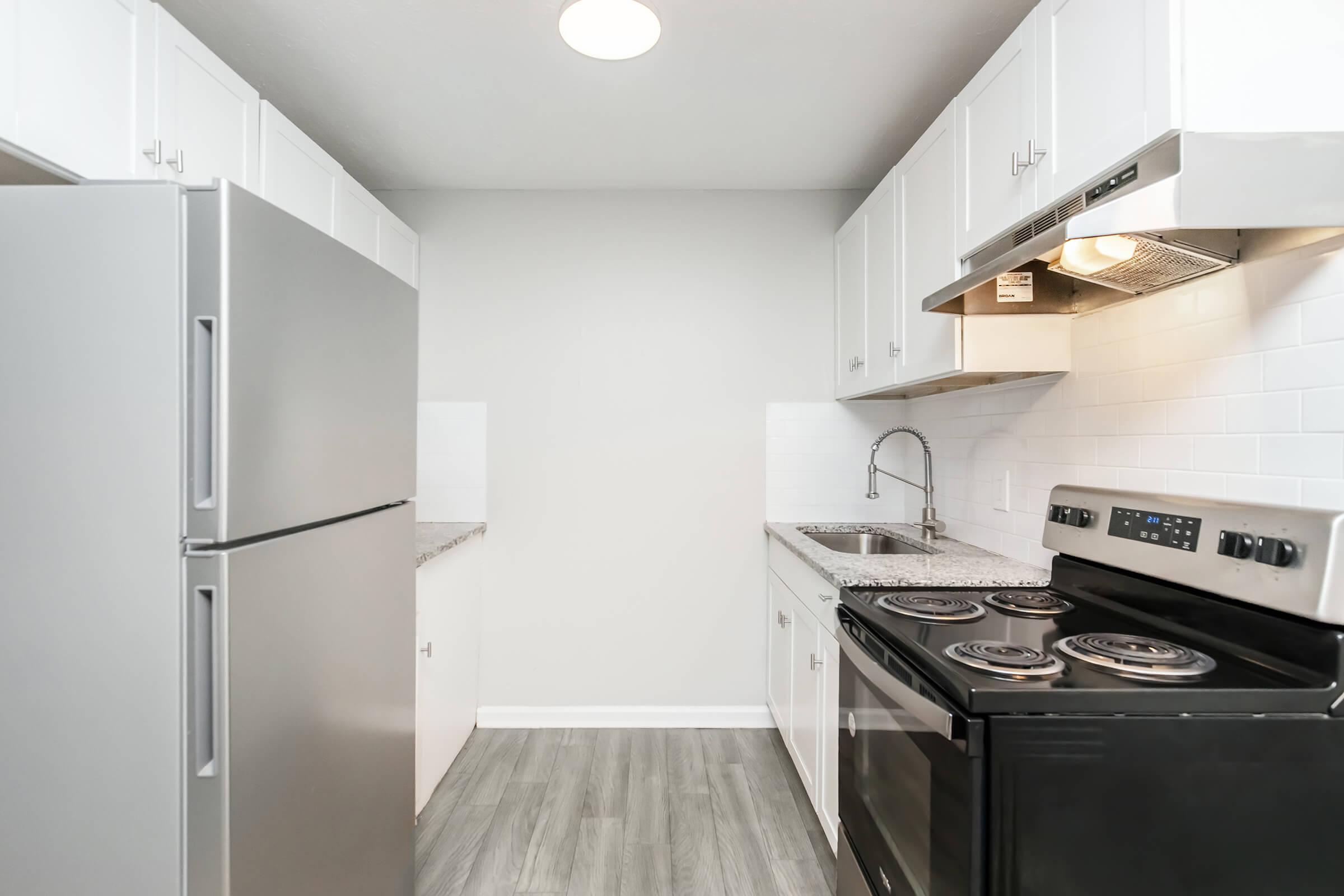 A modern kitchen featuring a stainless steel refrigerator, black stove, and white cabinetry. The countertops are light gray, and there is a small sink and backsplash. Natural light from a ceiling fixture illuminates the space, which has a neutral color palette and laminate flooring.