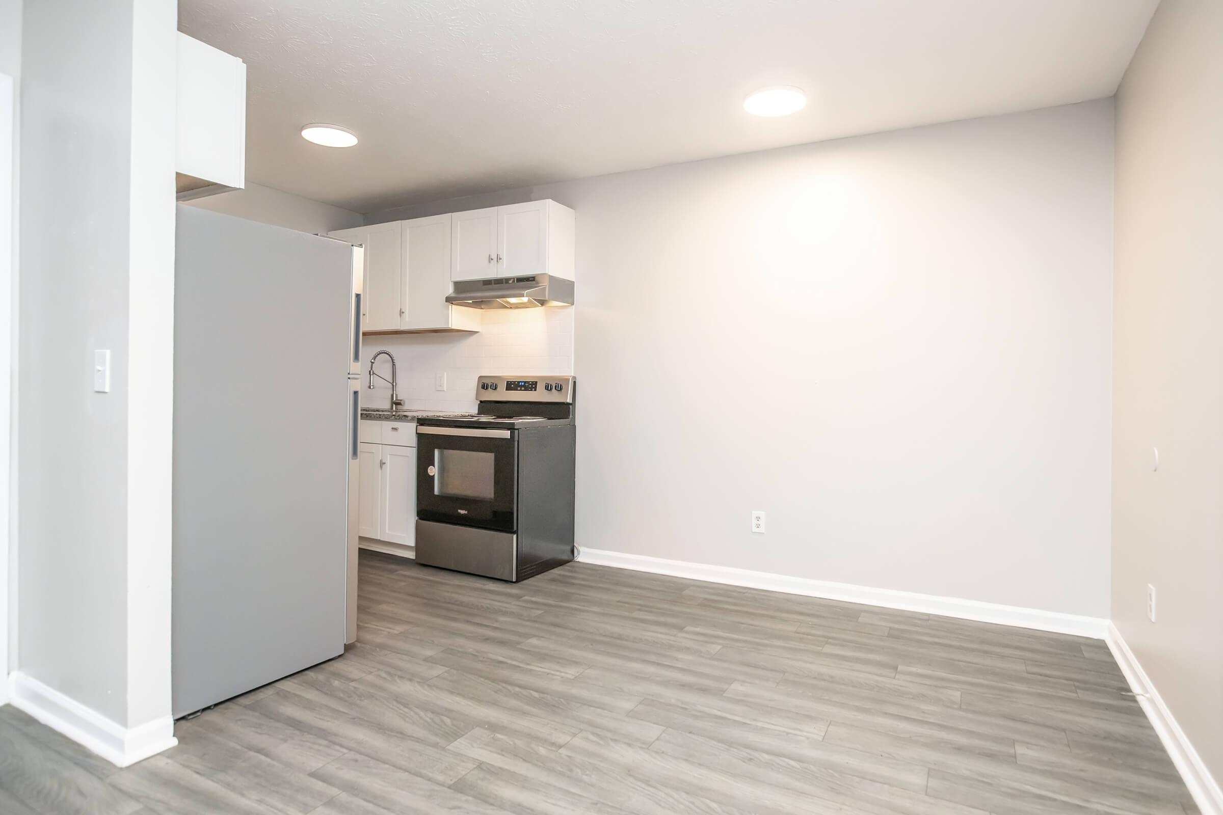 A modern kitchen space featuring a stainless steel refrigerator, an oven, and a hood over the stove. The walls are painted light gray, and the floor is a wood-like laminate. There is ample natural light from ceiling fixtures, with an open layout leading to an empty area, suggesting potential for dining or living space.