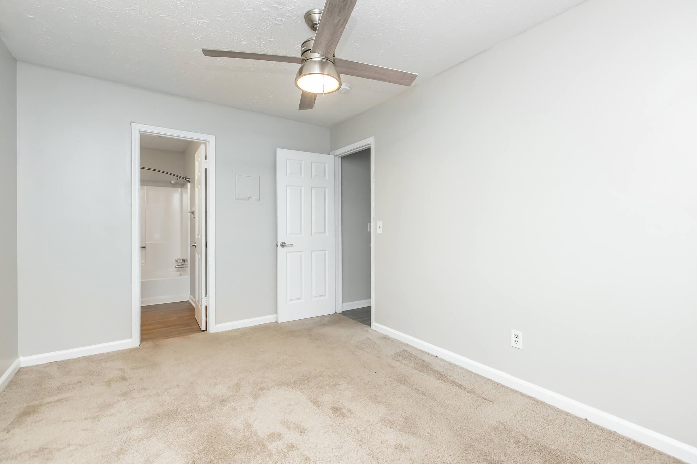 A spacious, empty room with light gray walls and plush beige carpet. The room features a modern ceiling fan and two doorways leading to adjacent areas, one with a bathroom visible. Natural light highlights the clean, uncluttered space, ideal for personalization or staging.