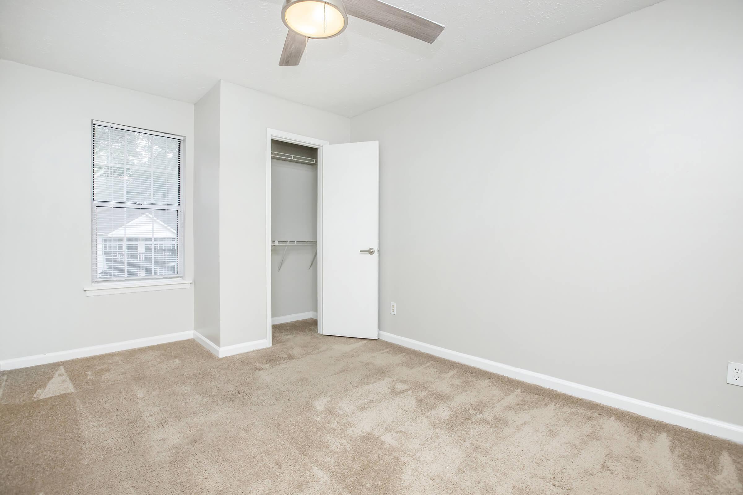 A neatly arranged bedroom with light gray walls and a light brown carpet. There is a closet with an open white door on the left. A ceiling fan hangs in the center of the room, and a window on the right allows natural light to enter. The overall atmosphere is clean and bright, perfect for a cozy living space.