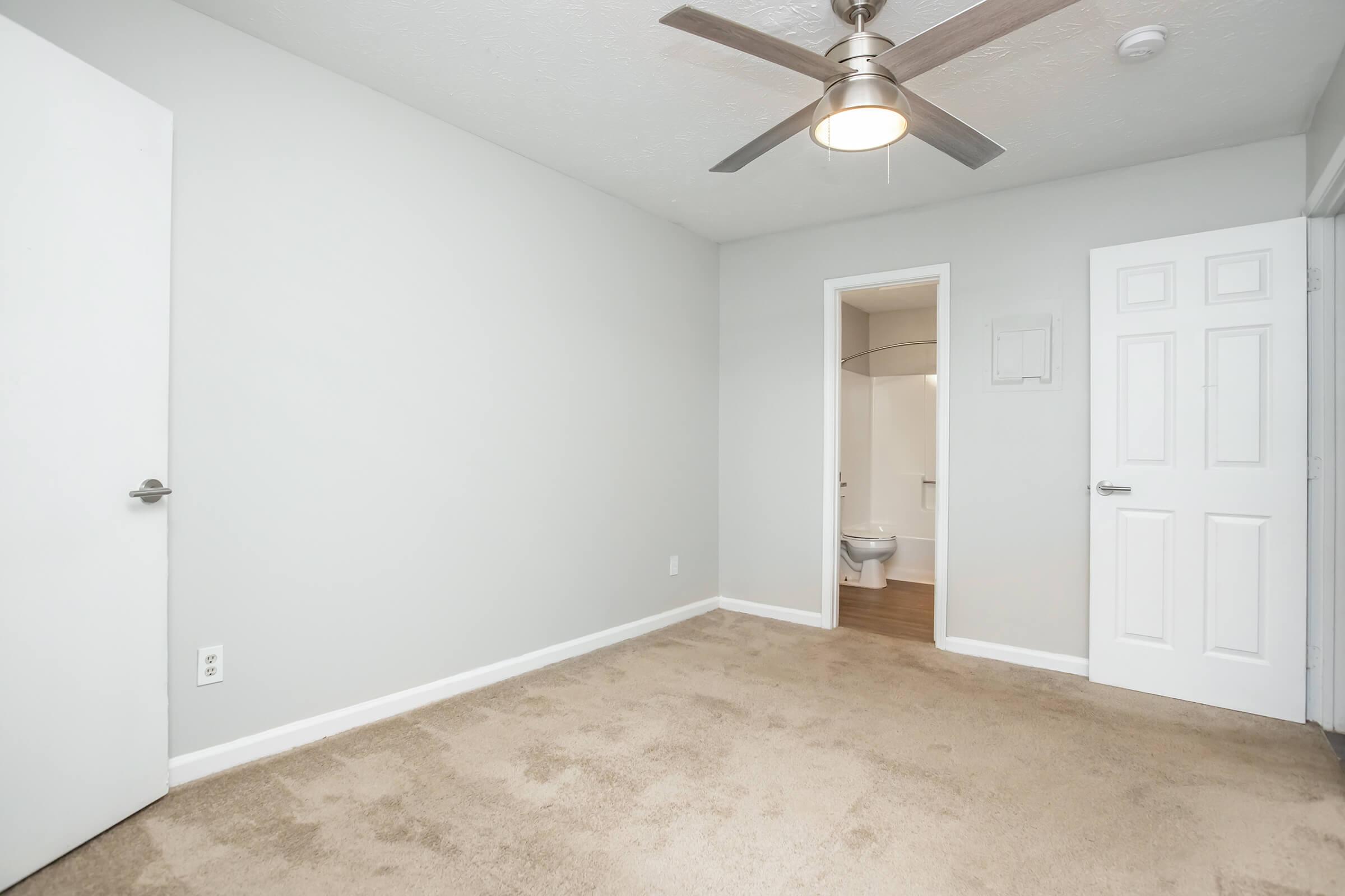 Empty room with light gray walls and beige carpet. A ceiling fan is visible, and there are two doors leading to adjacent areas, one of which appears to lead to a bathroom. Natural light enters the space, giving it a bright and open feel.