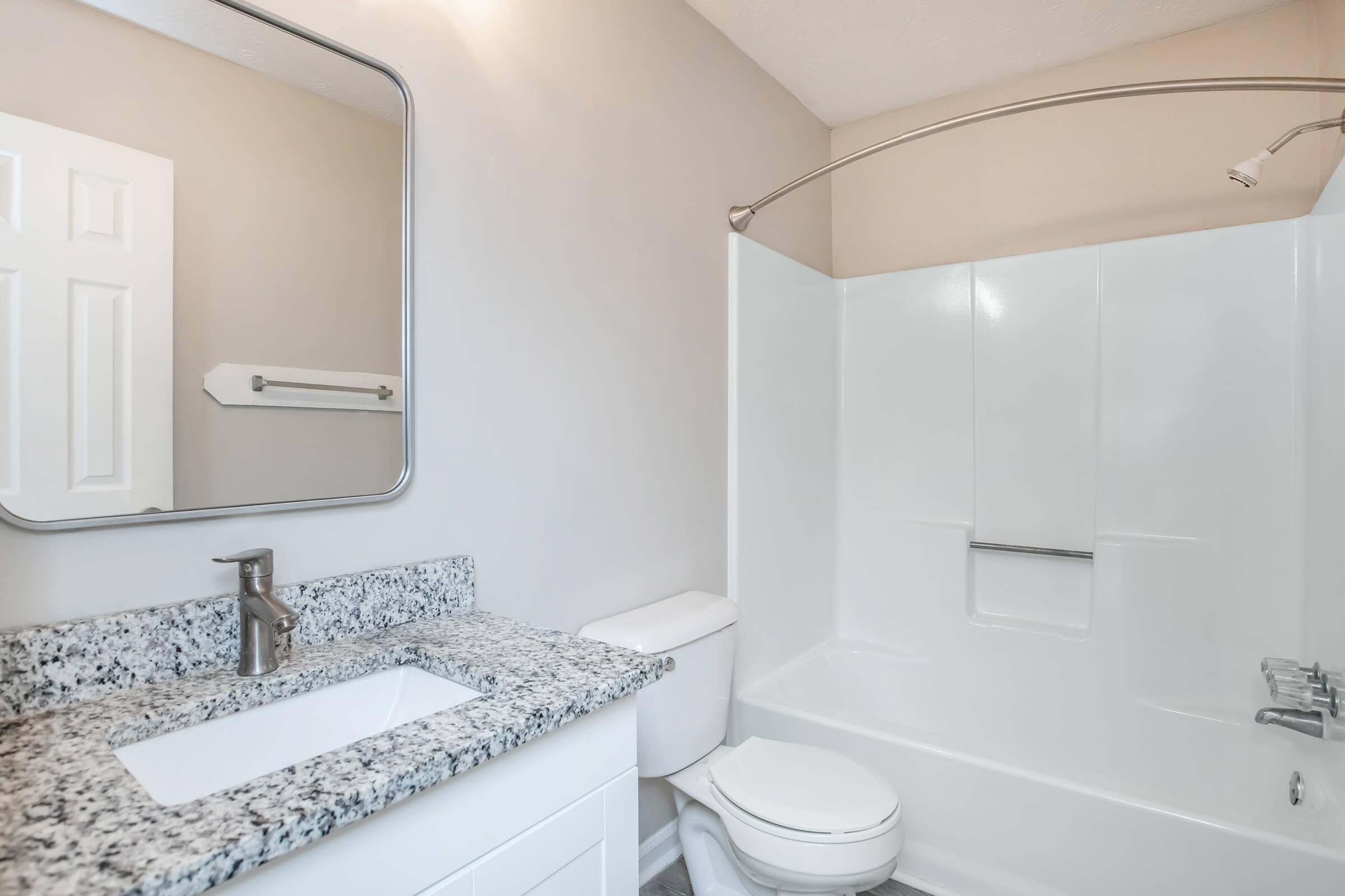 Modern bathroom featuring a white bathtub with a shower, a stylish granite countertop sink, and a mirror above the sink. The walls are painted light colors, and there's a towel rack near the door, enhancing the clean and minimalist design.