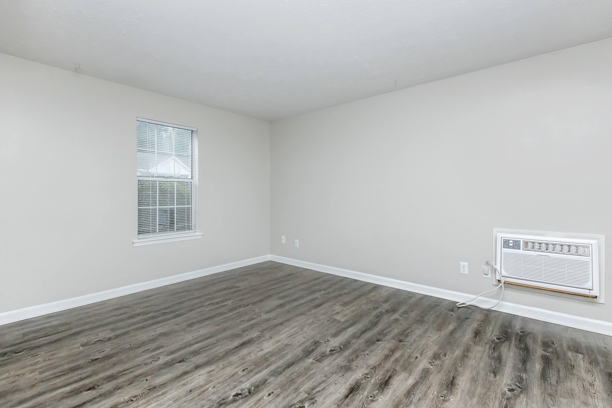 A simple, empty room with light gray walls and a window. The floor is covered in a wood-like laminate, and there's an air conditioning unit installed near the floor. The room has no furniture, emphasizing its spaciousness and minimalistic design.
