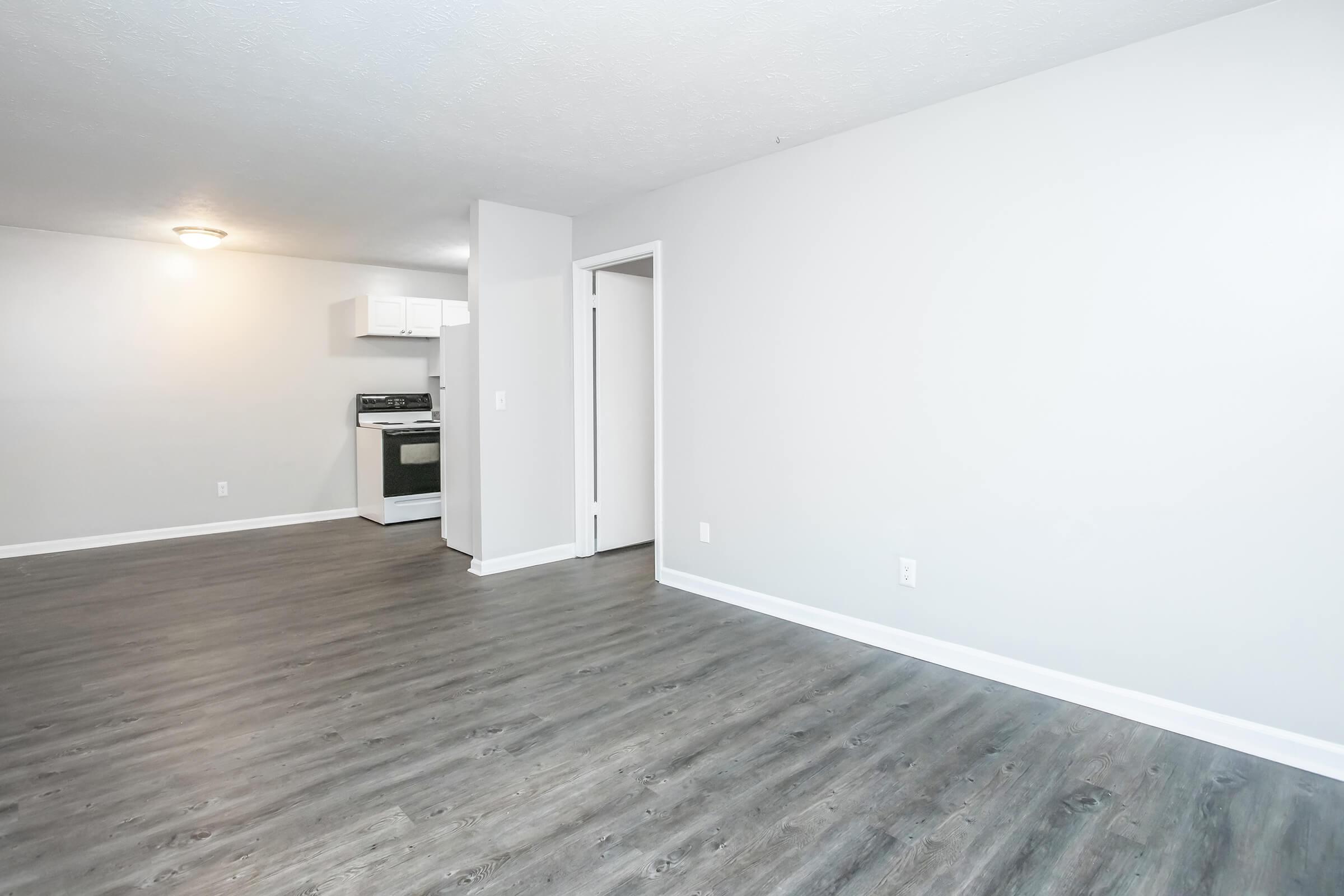 A spacious, vacant room with light gray walls and dark wood laminate flooring. In the background, a kitchen area is visible featuring a white refrigerator and an oven. A doorway to the right leads to another room, and a ceiling light casts illumination across the area.
